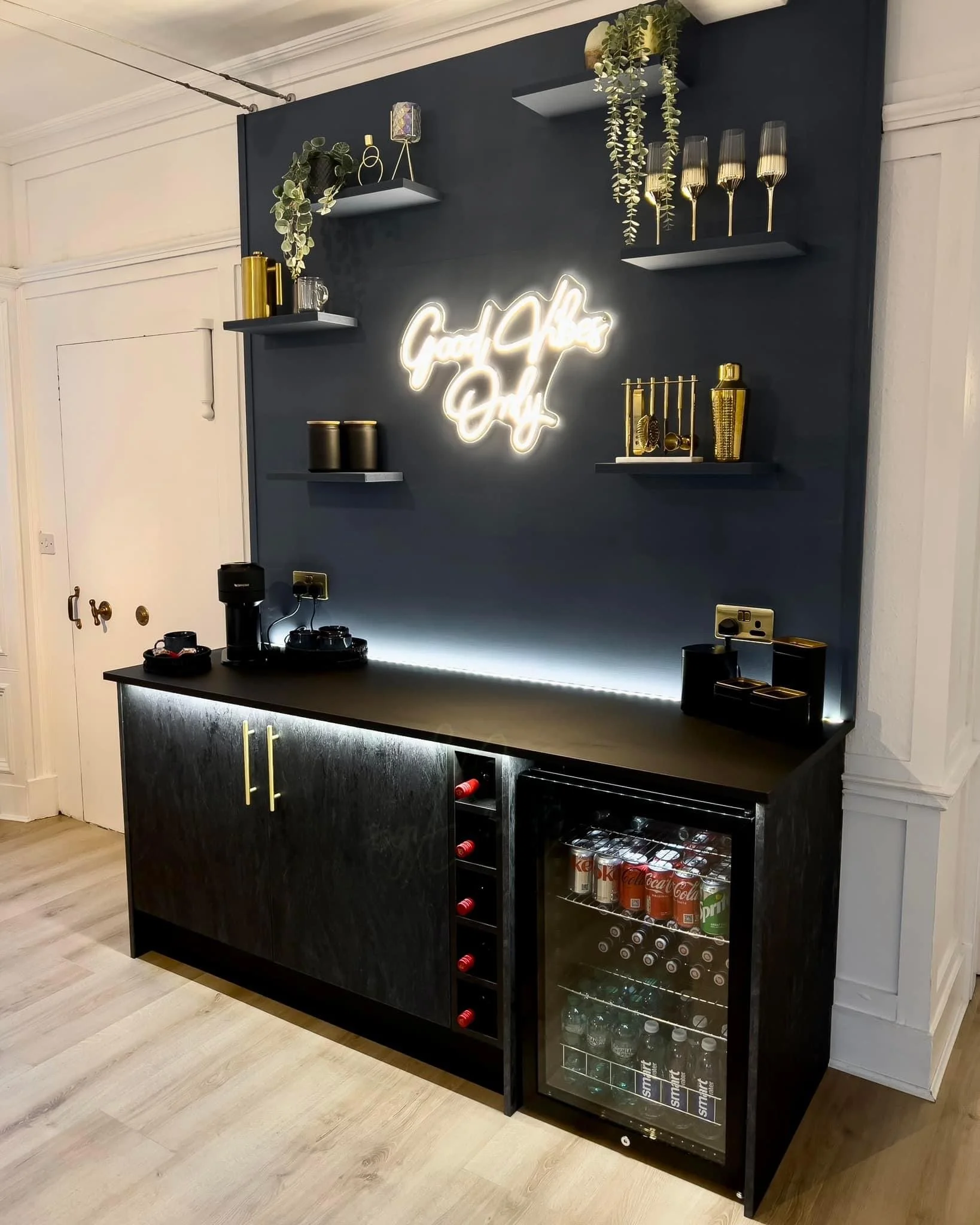 A modern beverage area with a black cabinet, a mini fridge filled with soft drinks, and a black countertop. Above the counter, a dark wall features floating shelves with decorative items, a neon sign that says "Good Vibes Only," and hanging wine glas