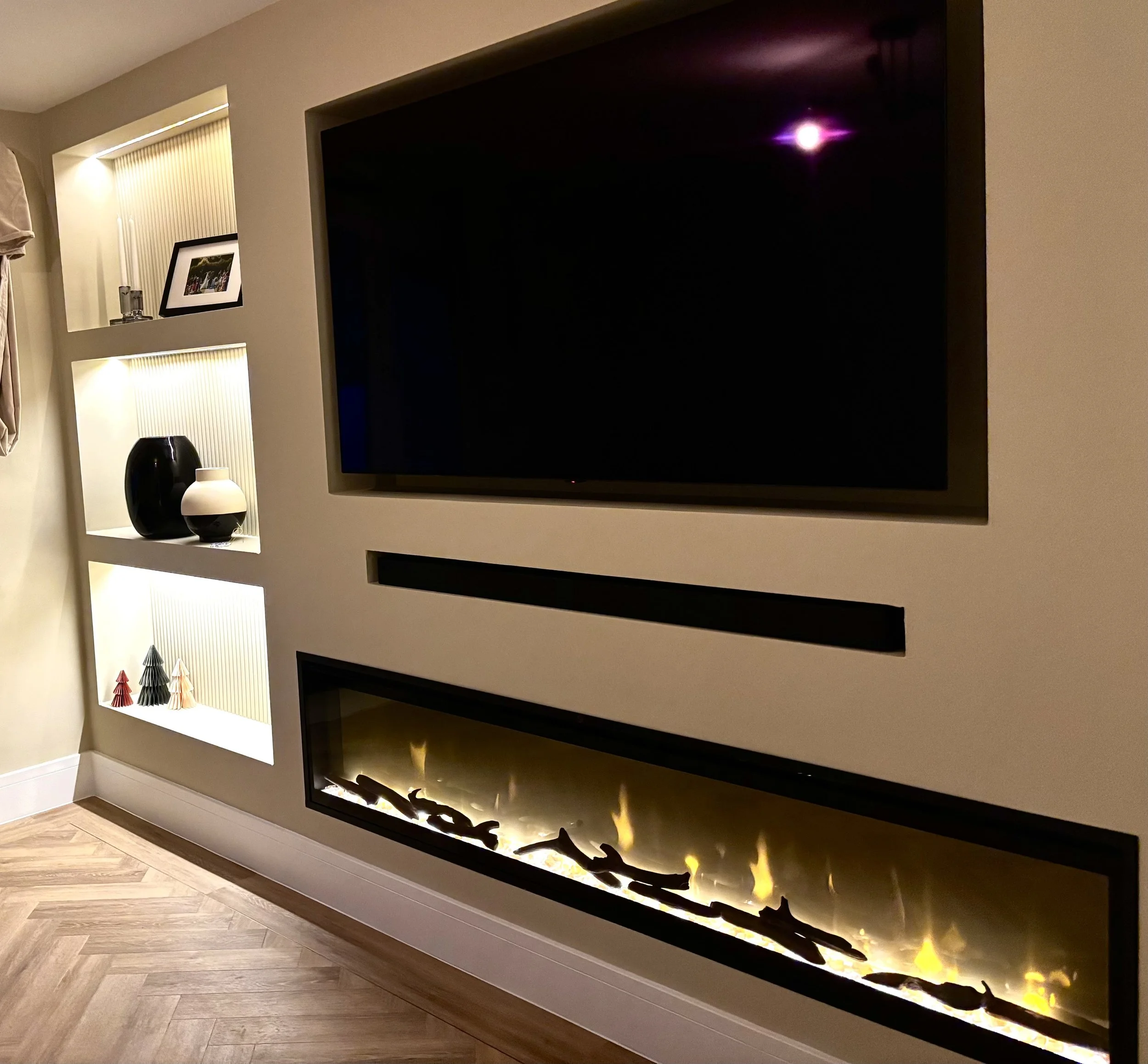 Living room with beige wall, built-in shelving with decorative vases and small Christmas trees, flat-screen TV, and modern gas fireplace.