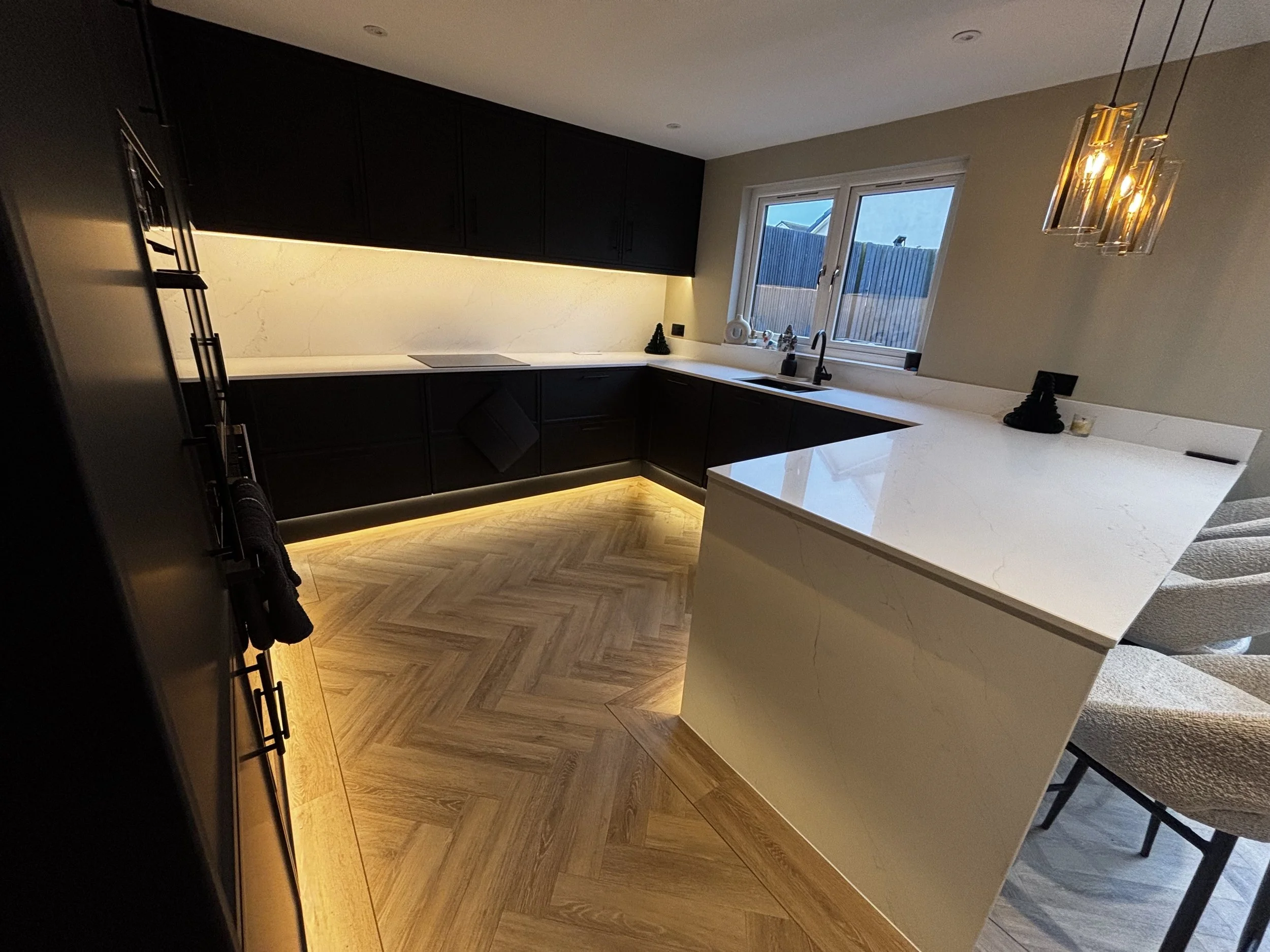 Modern kitchen with black cabinets, white marble countertops, herringbone wooden floor, and pendant lights