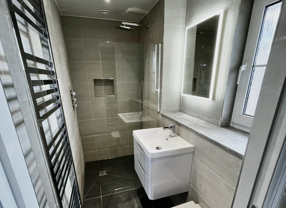 Modern bathroom with beige tiled walls, a walk-in shower with a rainfall showerhead, a built-in niche, a white vanity with a sink and faucet, a backlit mirror, and a window with a handle.