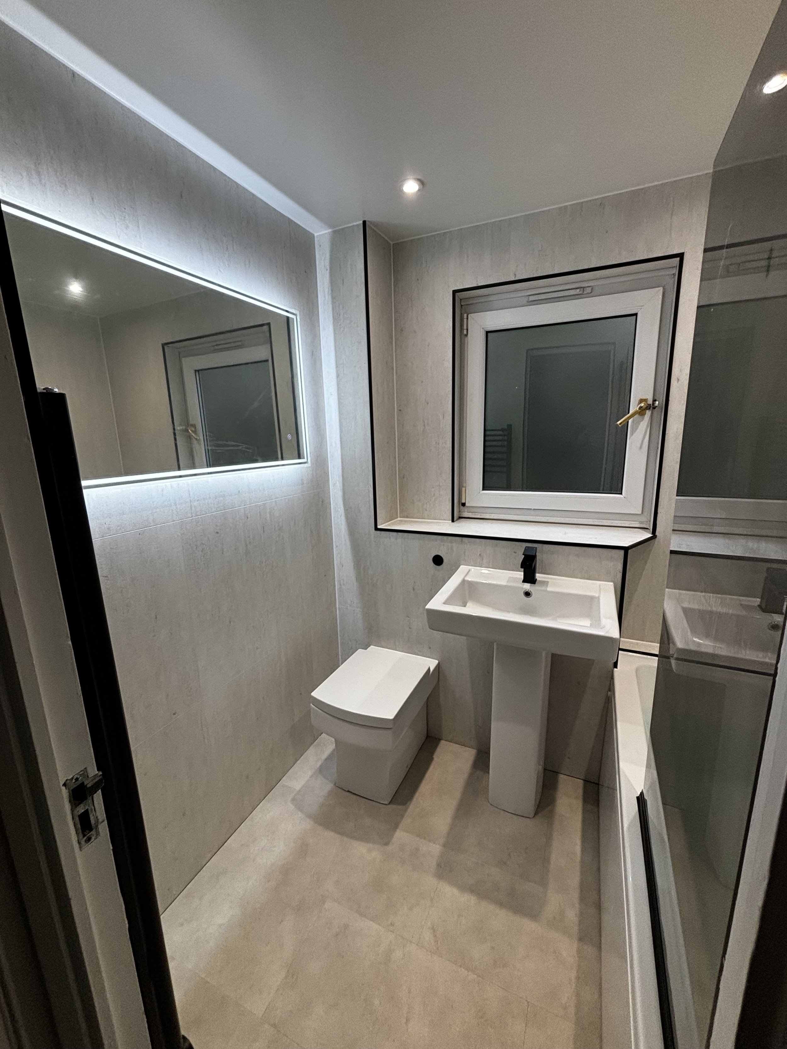Small bathroom with a toilet, a pedestal sink, a wall-mounted mirror with LED lighting, a window above the sink, and a glass shower enclosure.