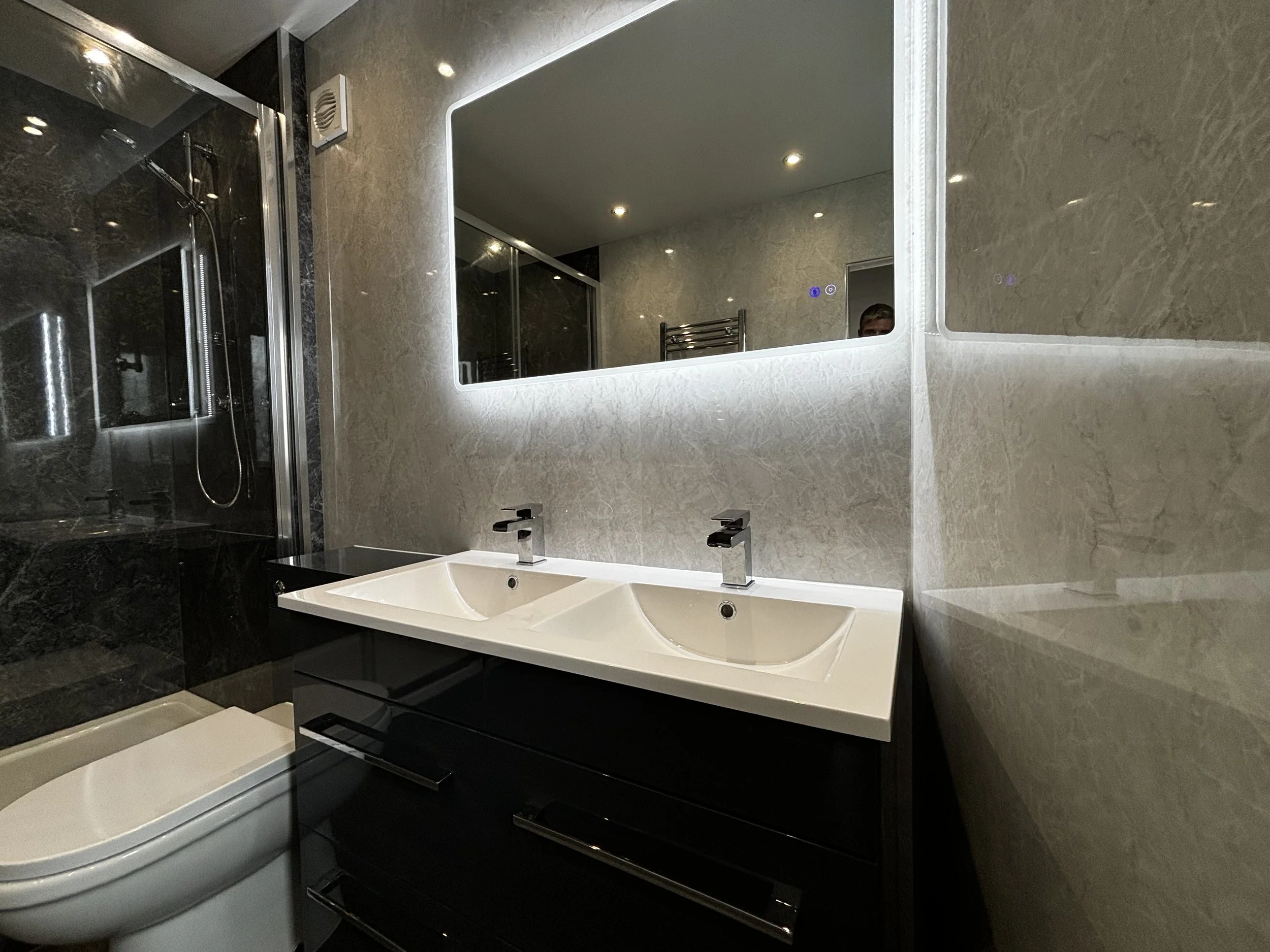 Modern bathroom with double sink vanity, large illuminated mirror, dark wood cabinet, and a walk-in shower with glass enclosure.