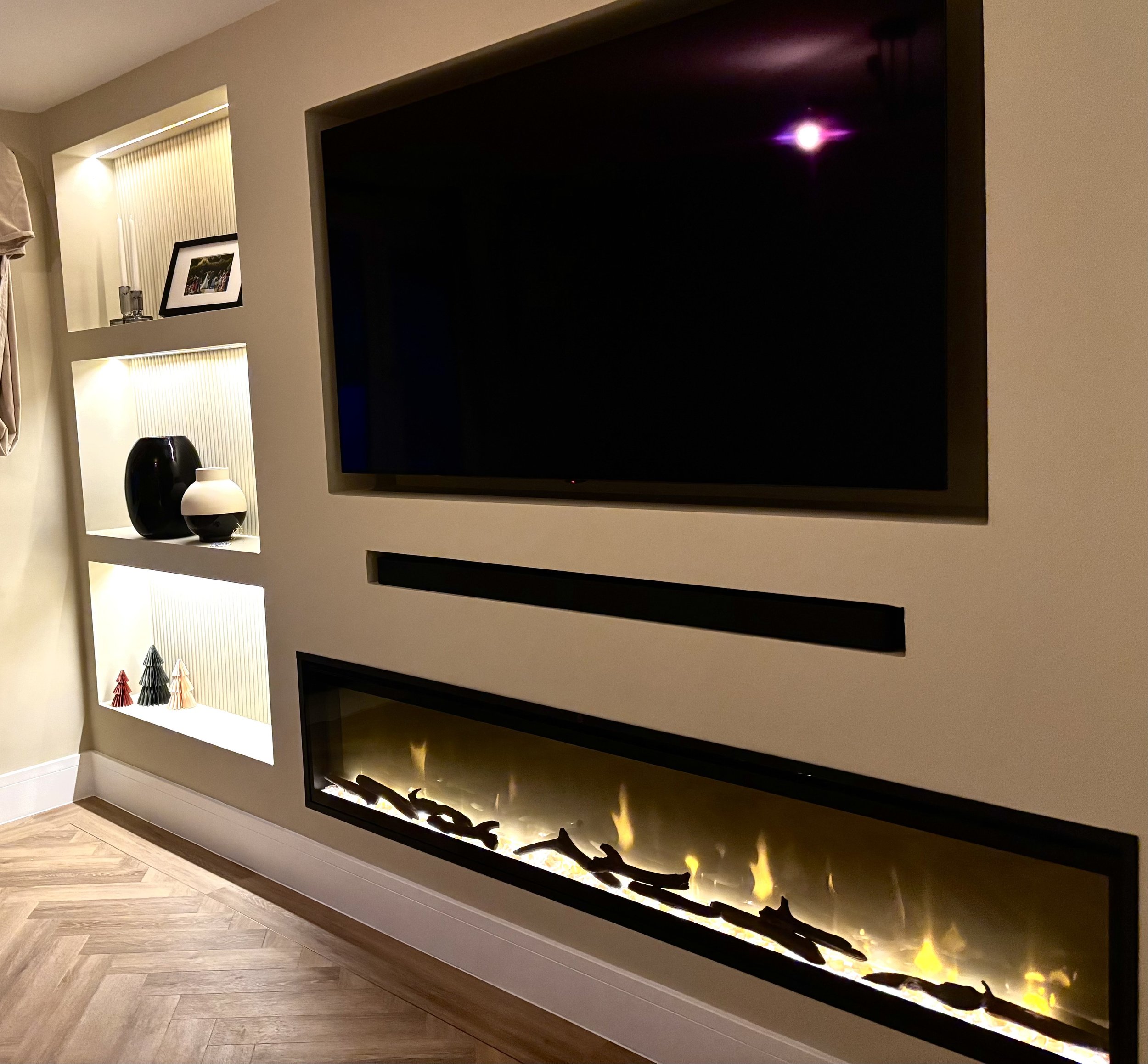 Living room wall with a large flat-screen TV, a modern fireplace below, and built-in shelves with decorative objects and framed photos.