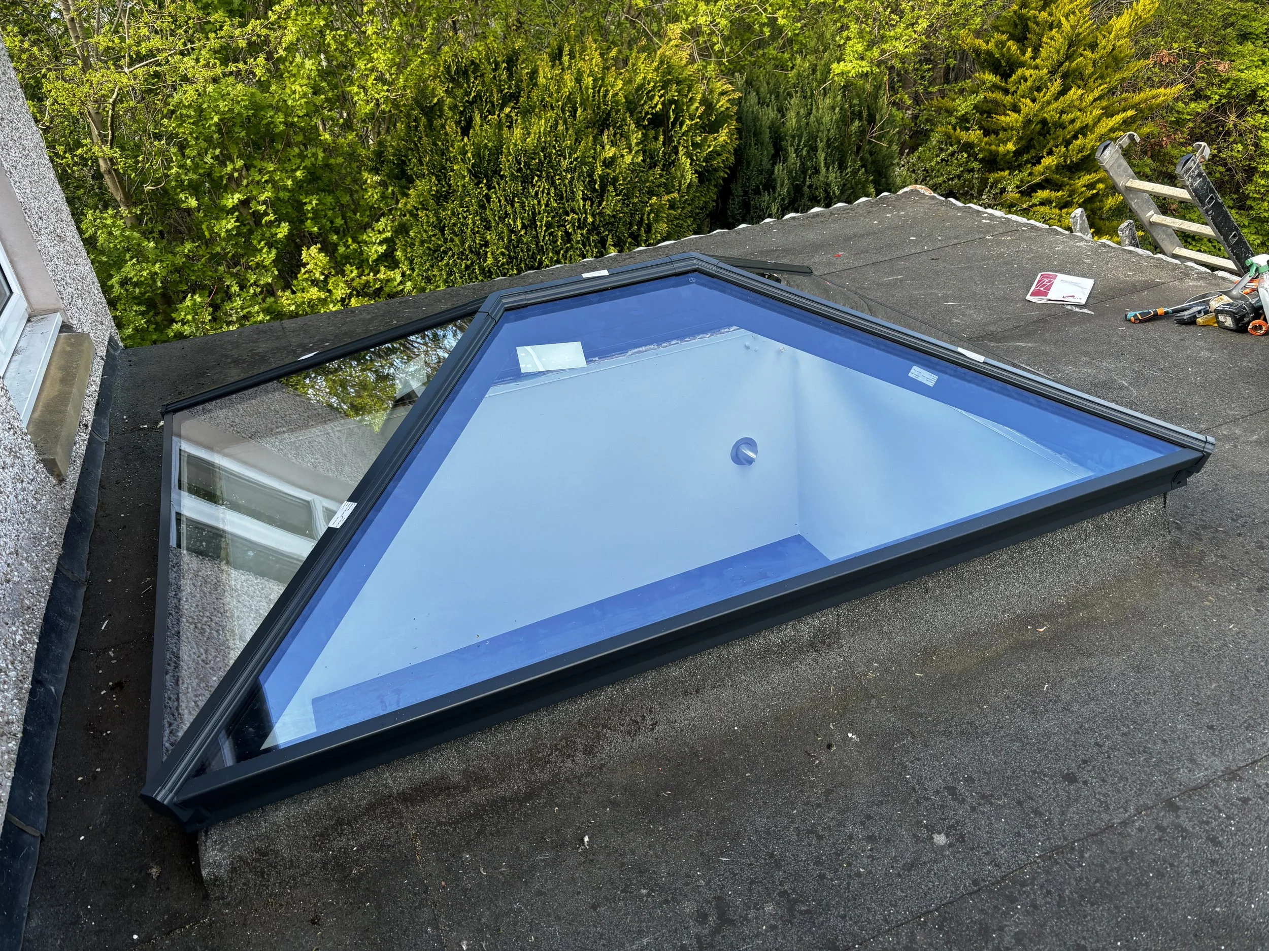 A skylight window installed on a flat roof, with tools and a ladder visible, overlooking green trees.