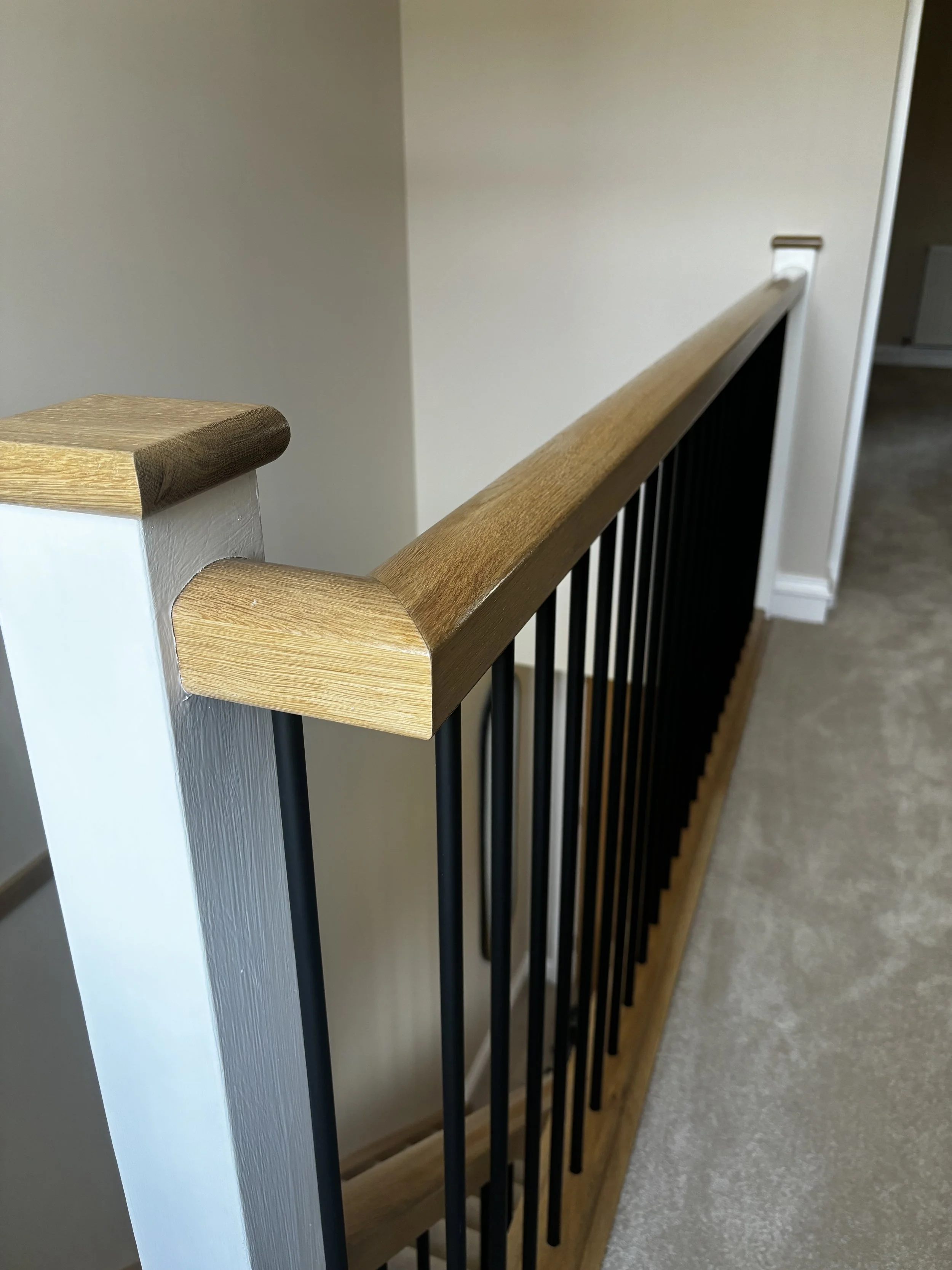 Close-up view of a wooden handrail and black metal balusters on a staircase or balcony, with a beige wall and carpeted floor in the background.