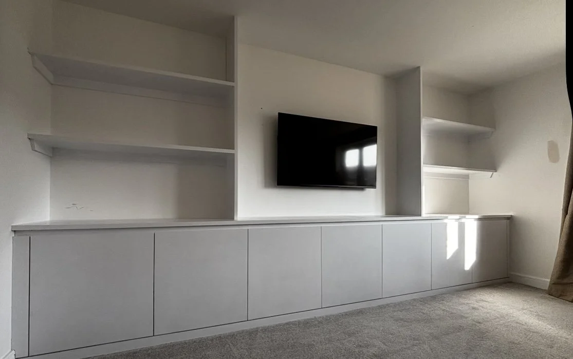 Empty white built-in entertainment unit with shelves and a wall-mounted television in a room with carpeted floor.