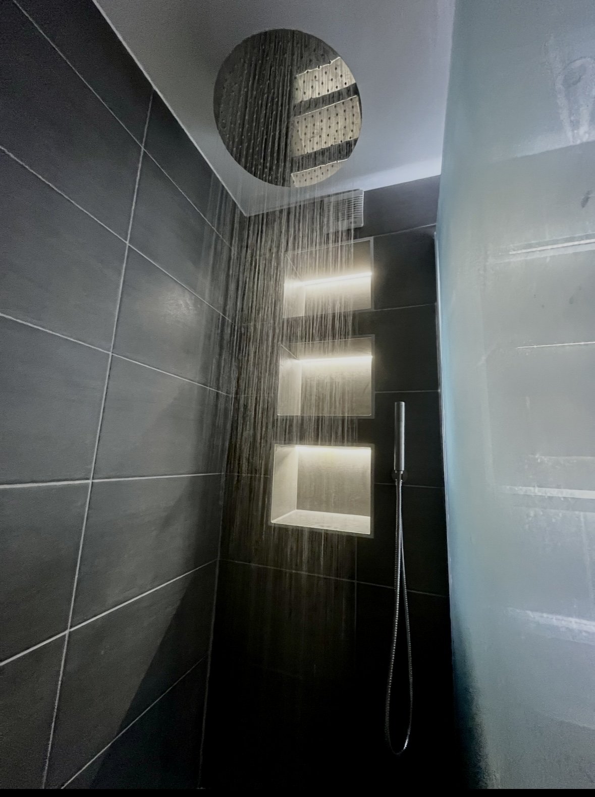 Close-up view of a modern shower with a round rainfall showerhead overhead, dark gray tiled walls, built-in rectangular shelves with lighting, and a handheld shower wand.