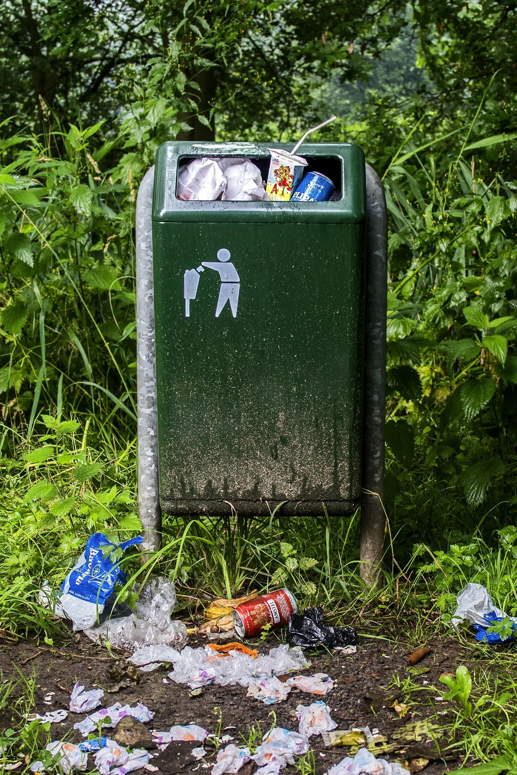 Une poubelle verte pour les déchets recyclables, remplie de déchets comme des papiers, une boisson et un emballage en plastique, entourée de déchets par terre dans un environnement naturel forestier.