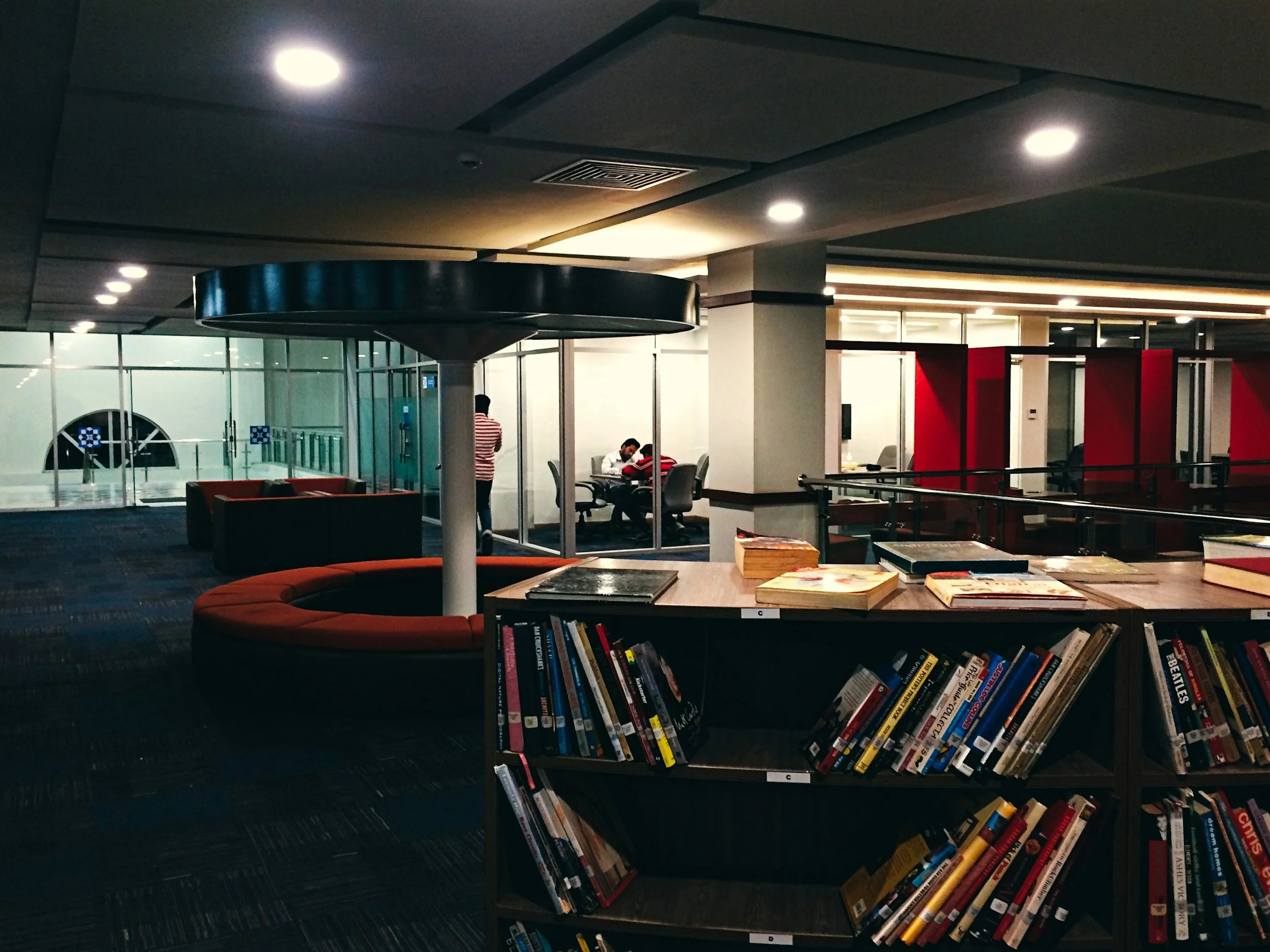 Espace de lecture dans un centre commercial ou une bibliothèque, avec une rangée de livres, des canapés rouges, des tables d'étude et des personnes assises ou debout.