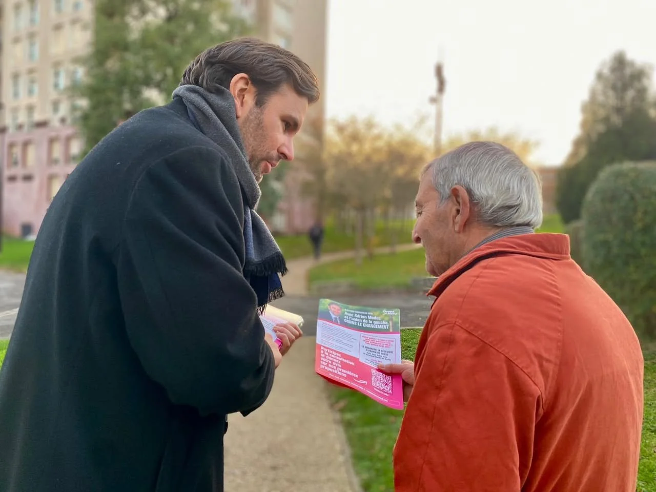 Deux hommes discutent à l'extérieur dans un parc ou une rue. L'un d'eux tient un prospectus rose. Il y a des arbres et des bâtiments en arrière-plan, avec une lumière douce.