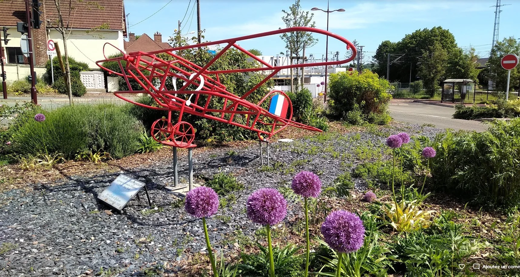 Une sculpture de petite avion rouge dans un jardin entouré de fleurs violettes et de végétation. En arrière-plan, il y a une rue avec des panneaux de signalisation.