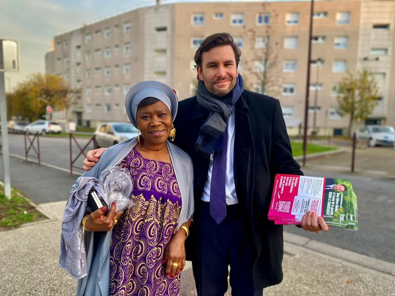 Deux personnes se tiennent côte à côte dans une rue. La femme porte une robe violette avec des motifs dorés et un manteau gris, ainsi qu'un foulard bleu. L'homme porte un manteau noir, une chemise blanche, une cravate violette et une écharpe grise. Il tient des brochures roses et vertes, l'homme a un sourire et pose avec la femme pour la photo.