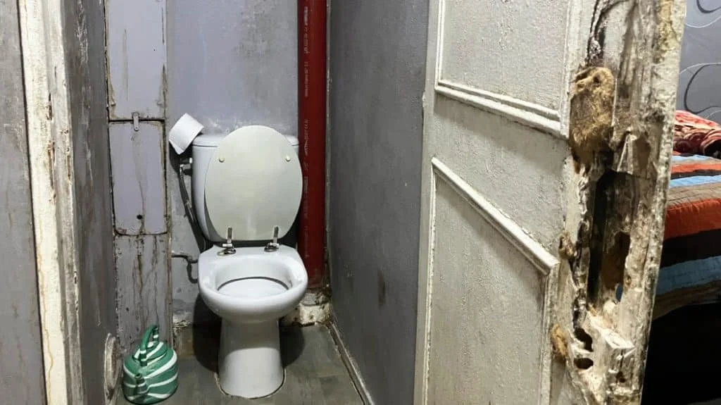 Toilettes dans une pièce en mauvais état avec une porte endommagée, murs en pierre et cartons de rangement à côté.