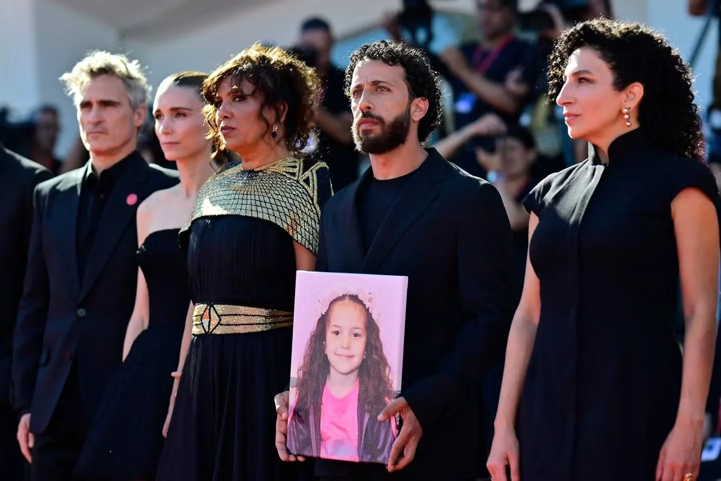 Groupe de cinq personnes en vêtement noir, debout lors d'une cérémonie ou un hommage. La personne au centre tient une photo d'une jeune fille avec des cheveux bouclés et un sourire.