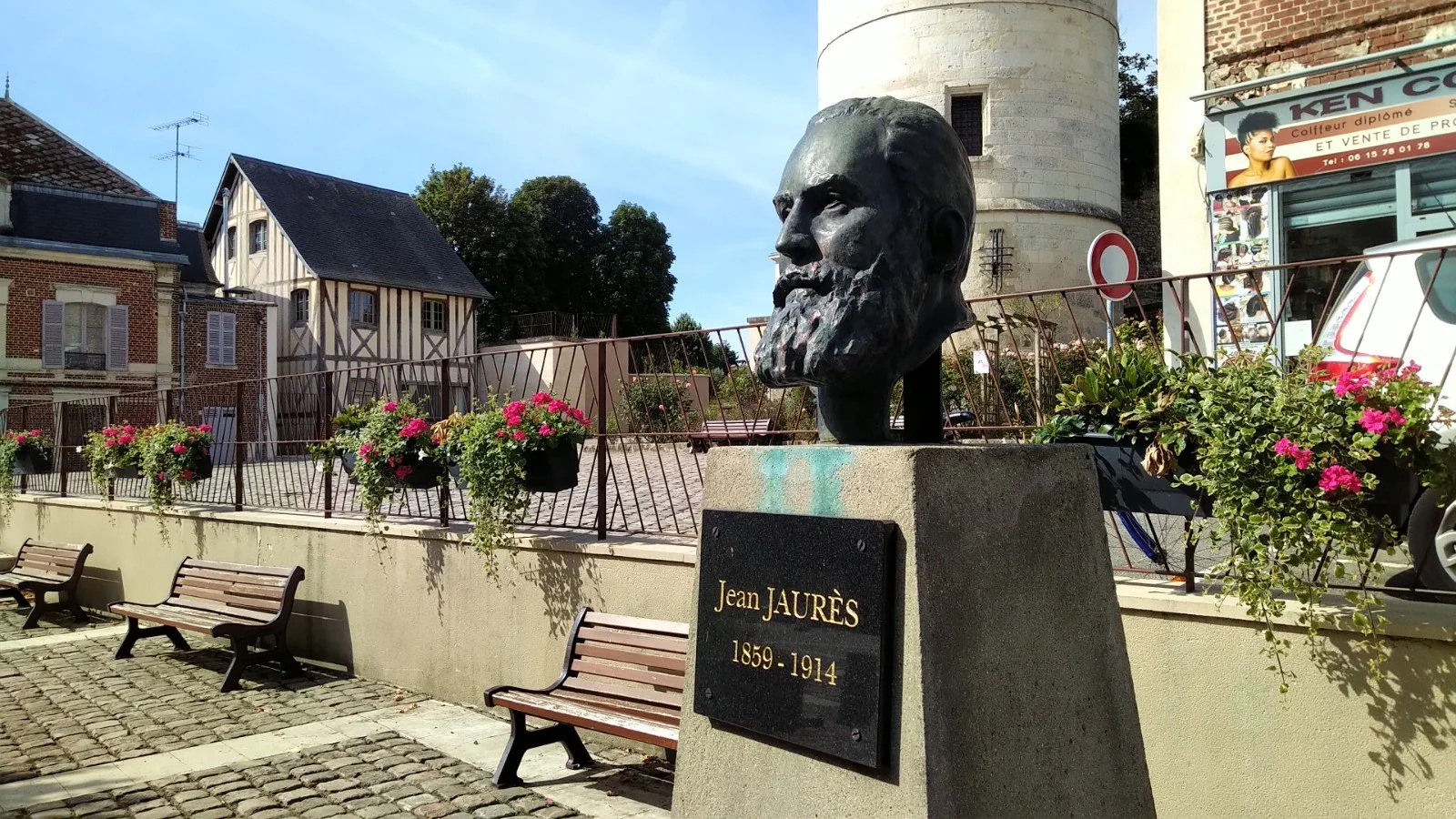 Statue en bronze de Jean Jaurès avec une plaque commémorative, entourée de fleurs et de bancs dans un environnement urbain.