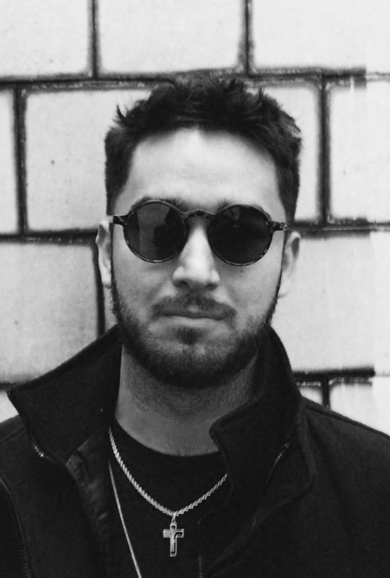 A black and white photo of a young man with dark hair, wearing round sunglasses, a black jacket, a chain necklace with a cross pendant, and a black shirt, standing against a brick wall.