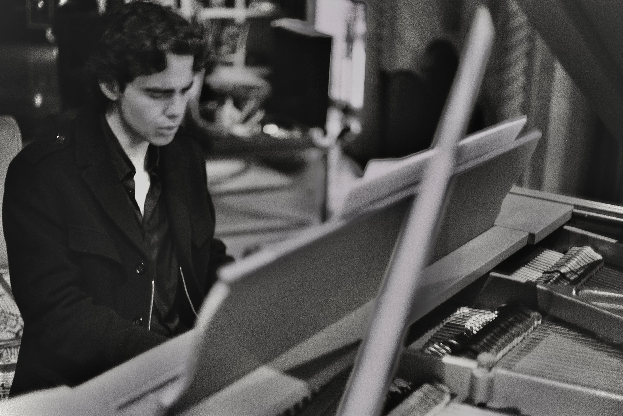 A woman playing the piano, seated at the keyboard with focus, in a room with music stands and other piano instruments visible in the background.
