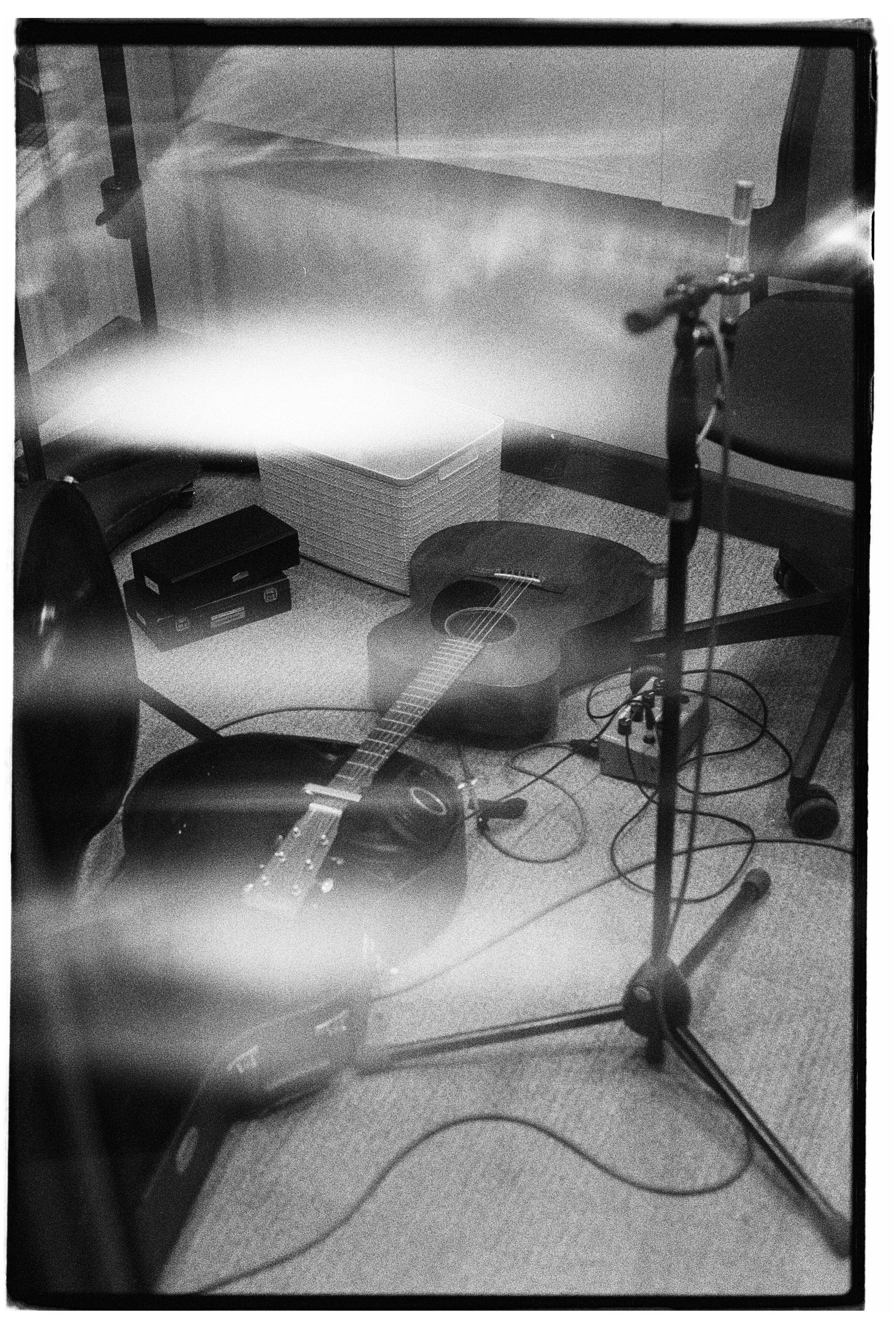 A black and white photo of a room with a guitar on the floor, a microphone stand, a chair, a small amplifier, a box of paper, and some cables.