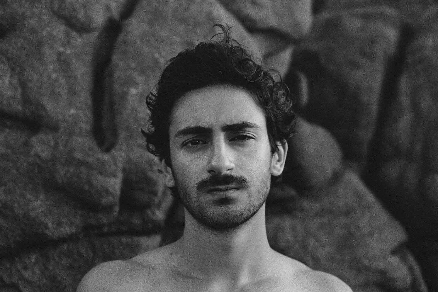 A black-and-white portrait of a young man with curly hair and a serious expression standing in front of a rocky background.