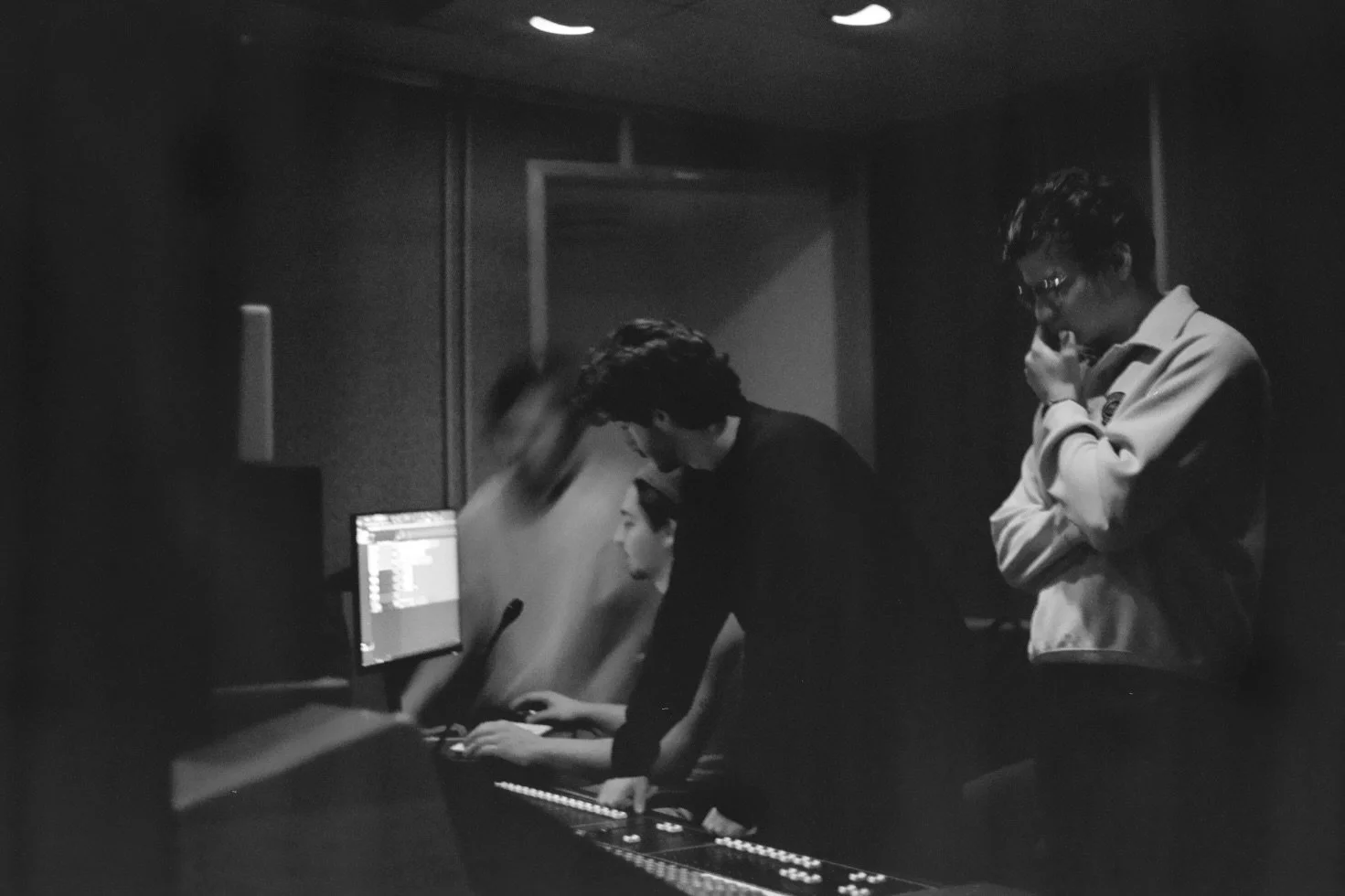 Black and white photo of three young people in a recording studio. One person is sitting at a mixing console, another is standing and leaning over a computer monitor, and a third person is standing with hand near mouth, possibly thinking or observing.