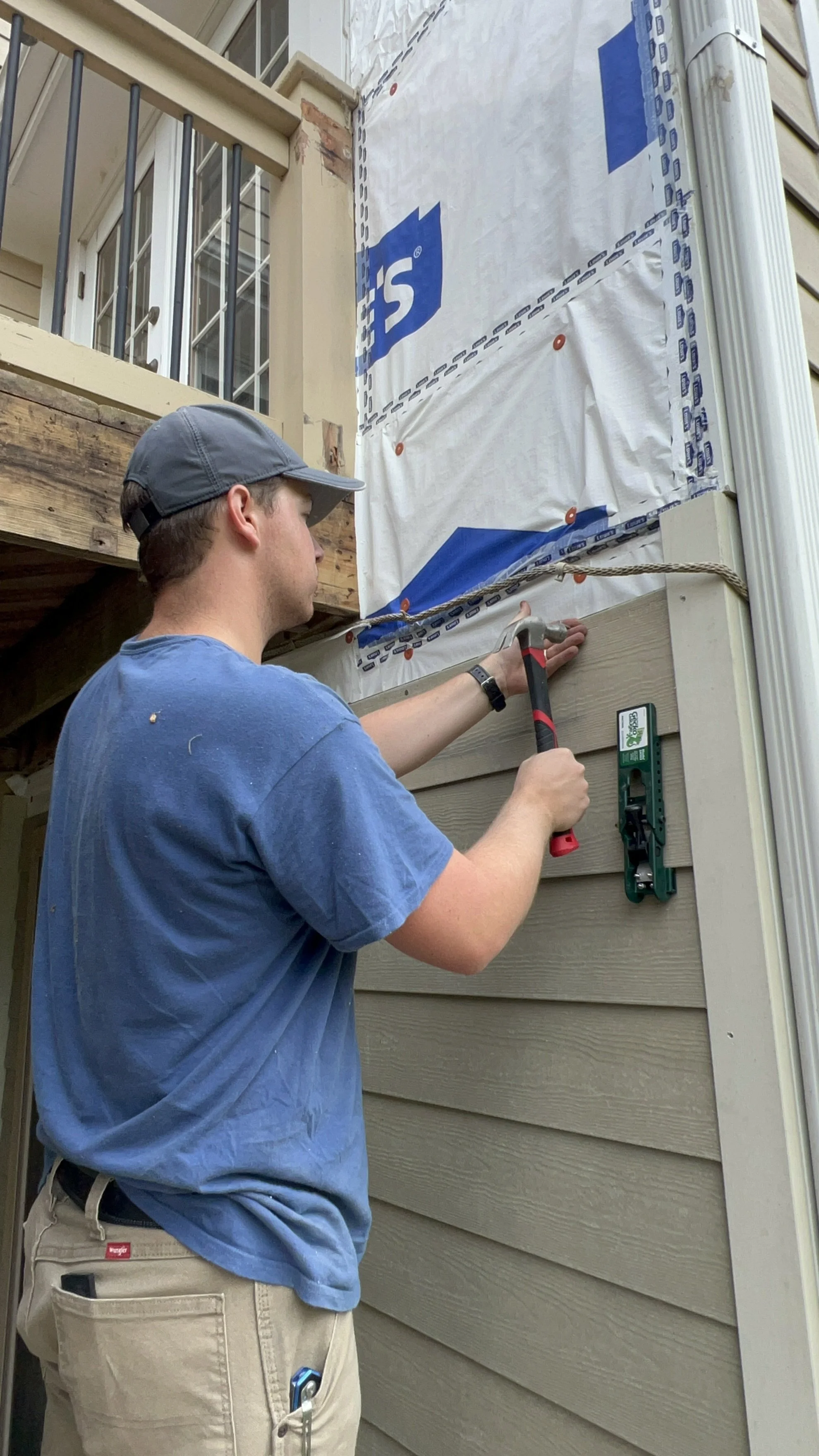 A man wearing a blue t-shirt, khaki shorts, and a gray cap is hammering a piece of siding onto the exterior wall of a house during construction or renovation. The house has beige horizontal siding and some construction material is visible in the background.