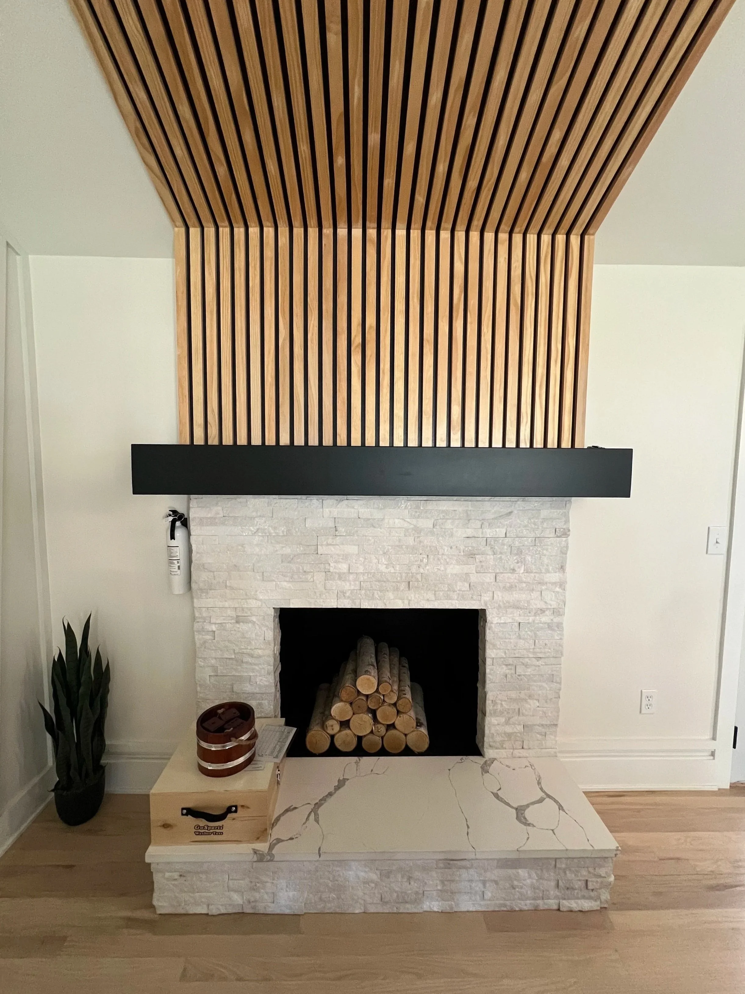 Modern fireplace with stacked logs, surrounded by white brick, with a black horizontal mantle and wooden slat ceiling and wall behind, in a minimalist living space.
