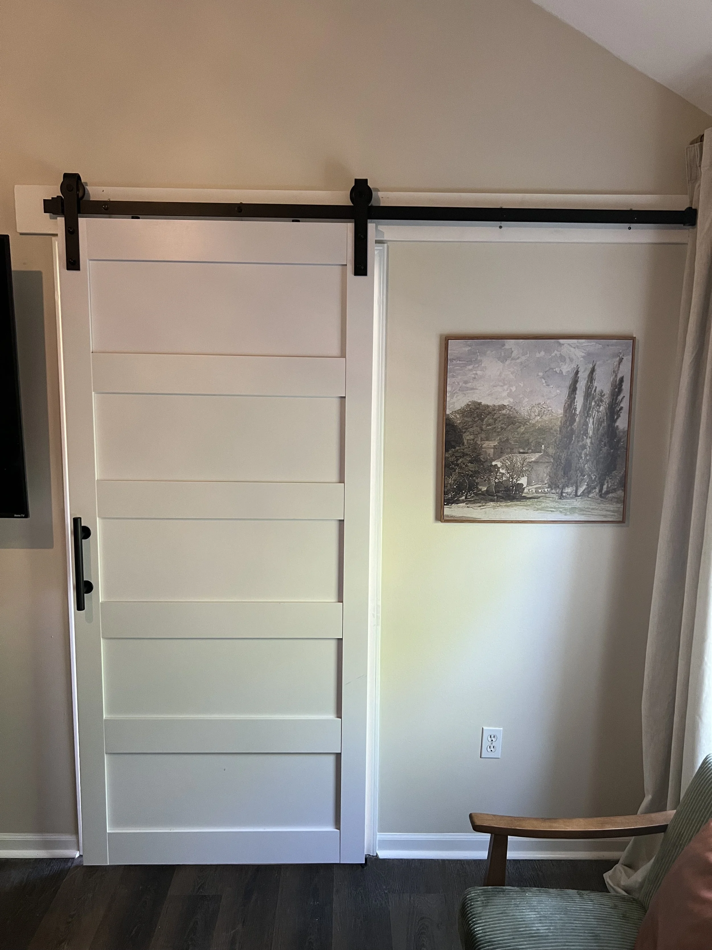 A white sliding barn door with black hardware installed on a wall next to a framed landscape painting and a window with curtains in a room.
