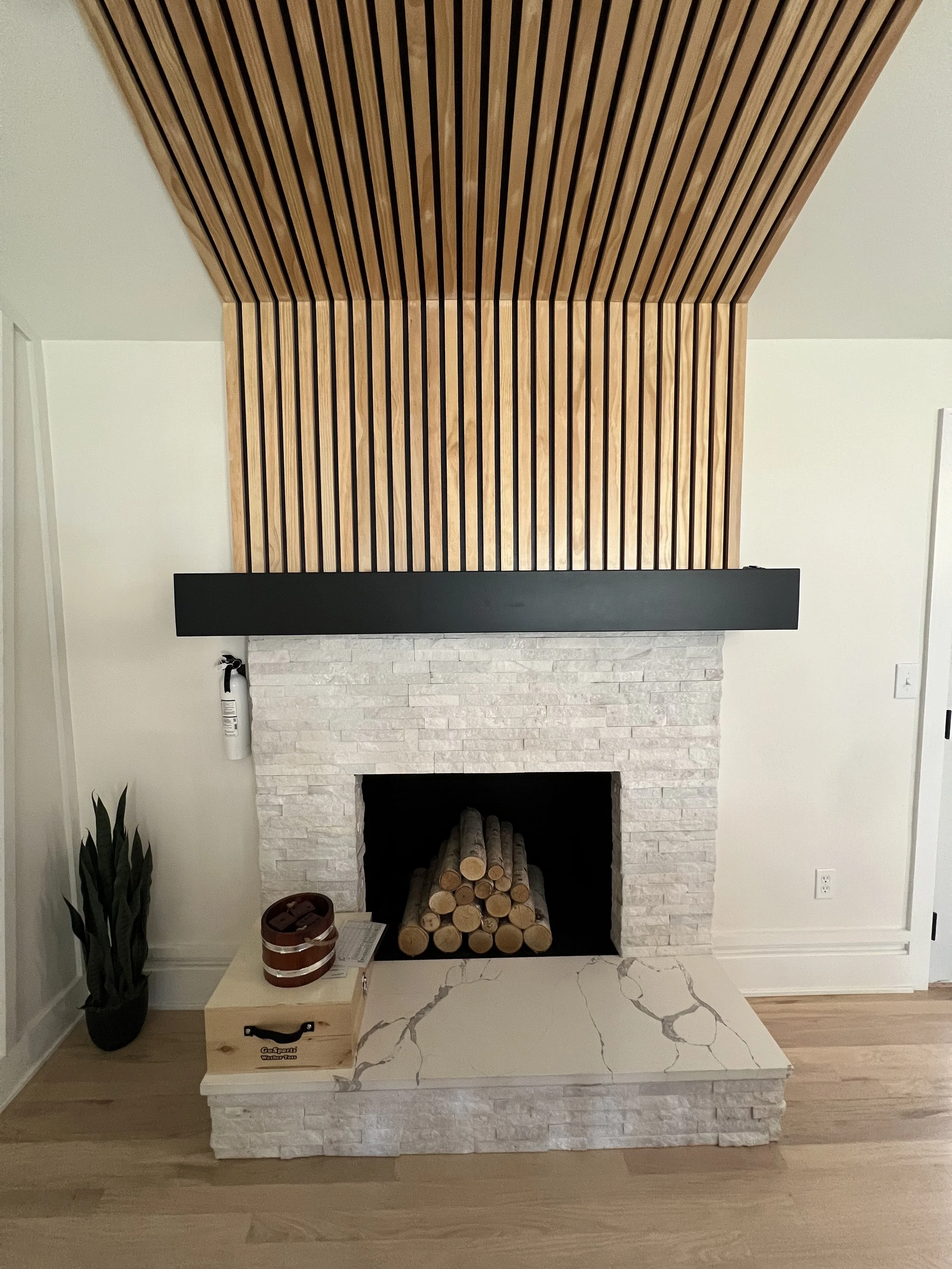 Modern living room fireplace with light-colored stone, wooden logs, and decorative plant on the side, topped with a wooden ceiling feature.