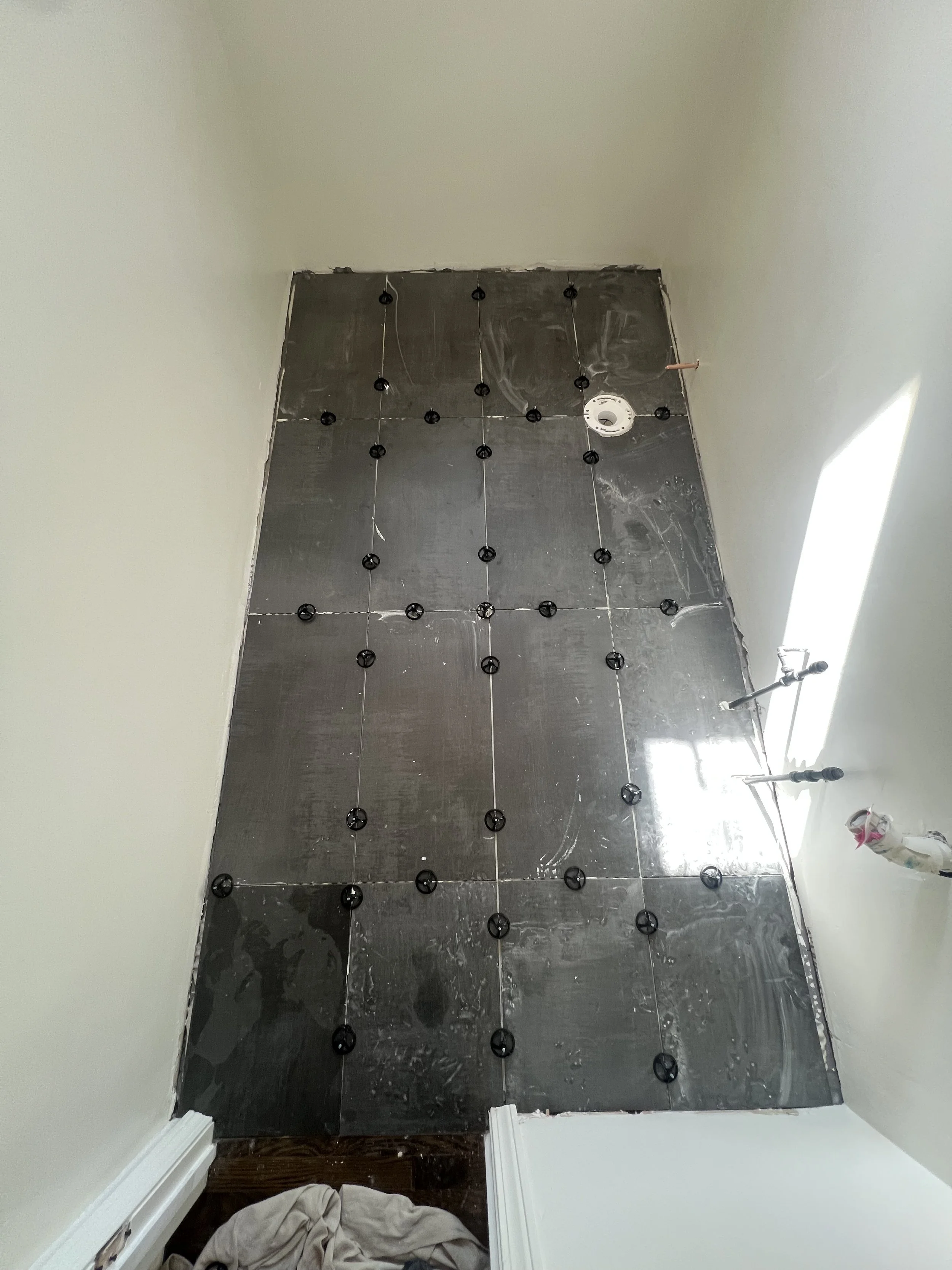 Bathroom floor under construction with black tiles and spacers, with some unfinished edges and plumbing pipes visible on the right side.