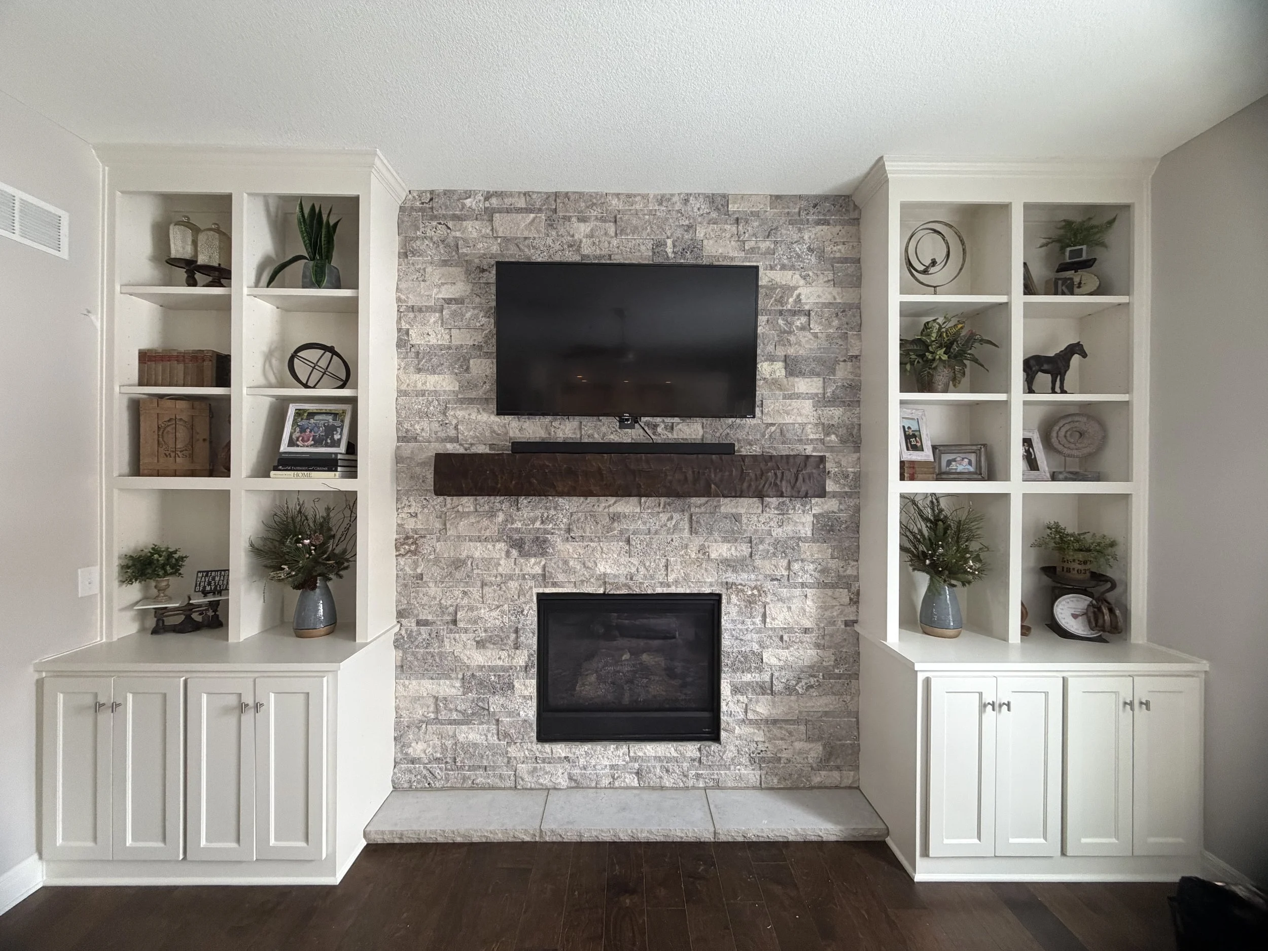 Living room with a stone fireplace, mounted TV, and built-in white cabinets on either side with decorative items, plants, and framed photos.