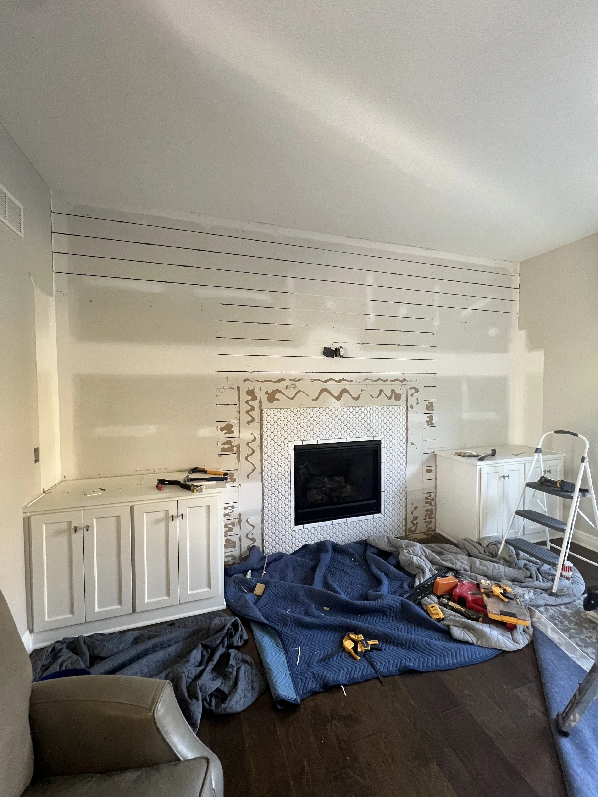 Living room under renovation with tools, step ladder, and blankets on the floor in front of a fireplace and wall paneling.