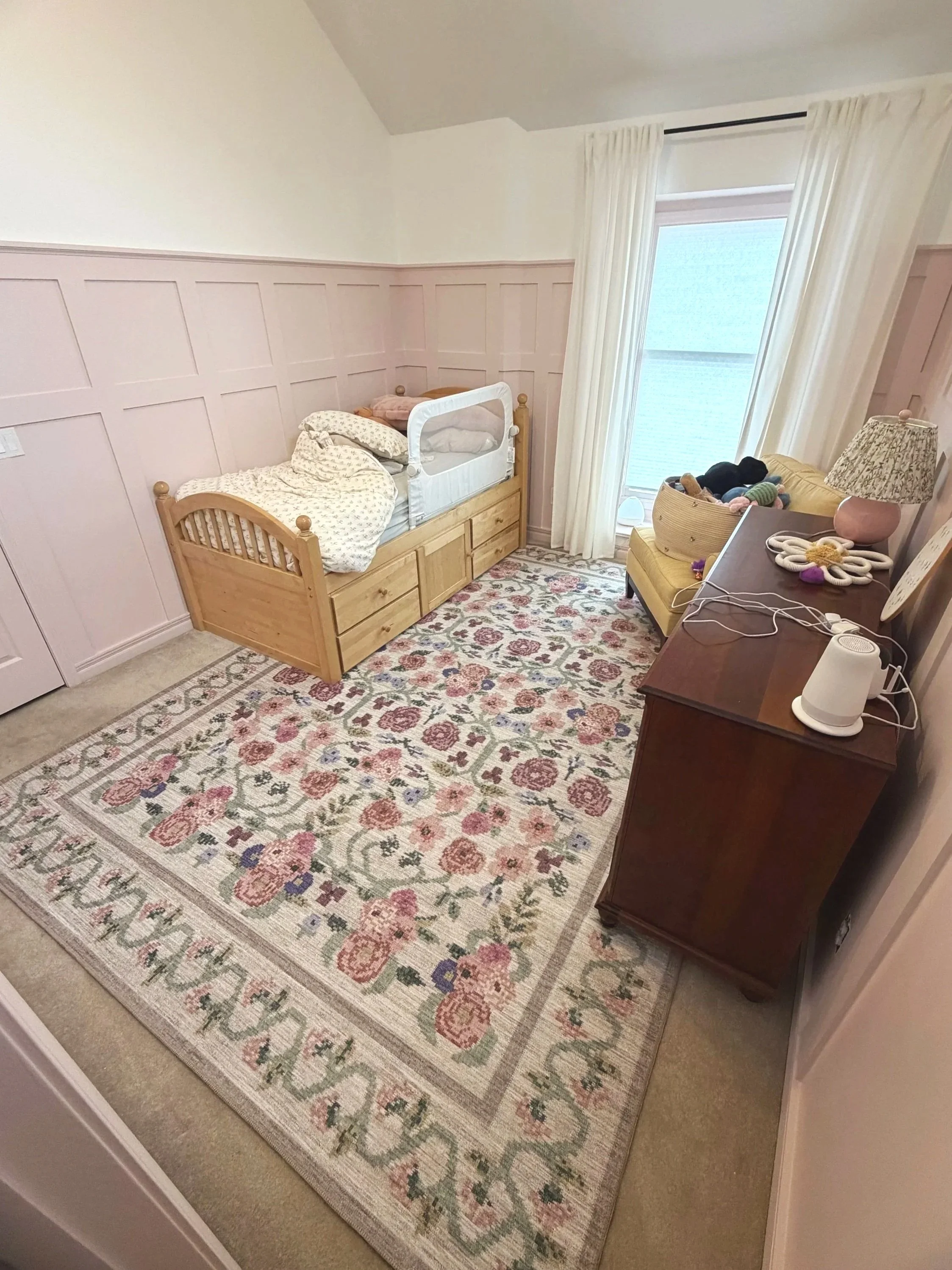 A cozy nursery with a wooden crib, a yellow armchair, a white dresser with a lamp, and a floral area rug. The room has pink wainscoting and a large window with white curtains.