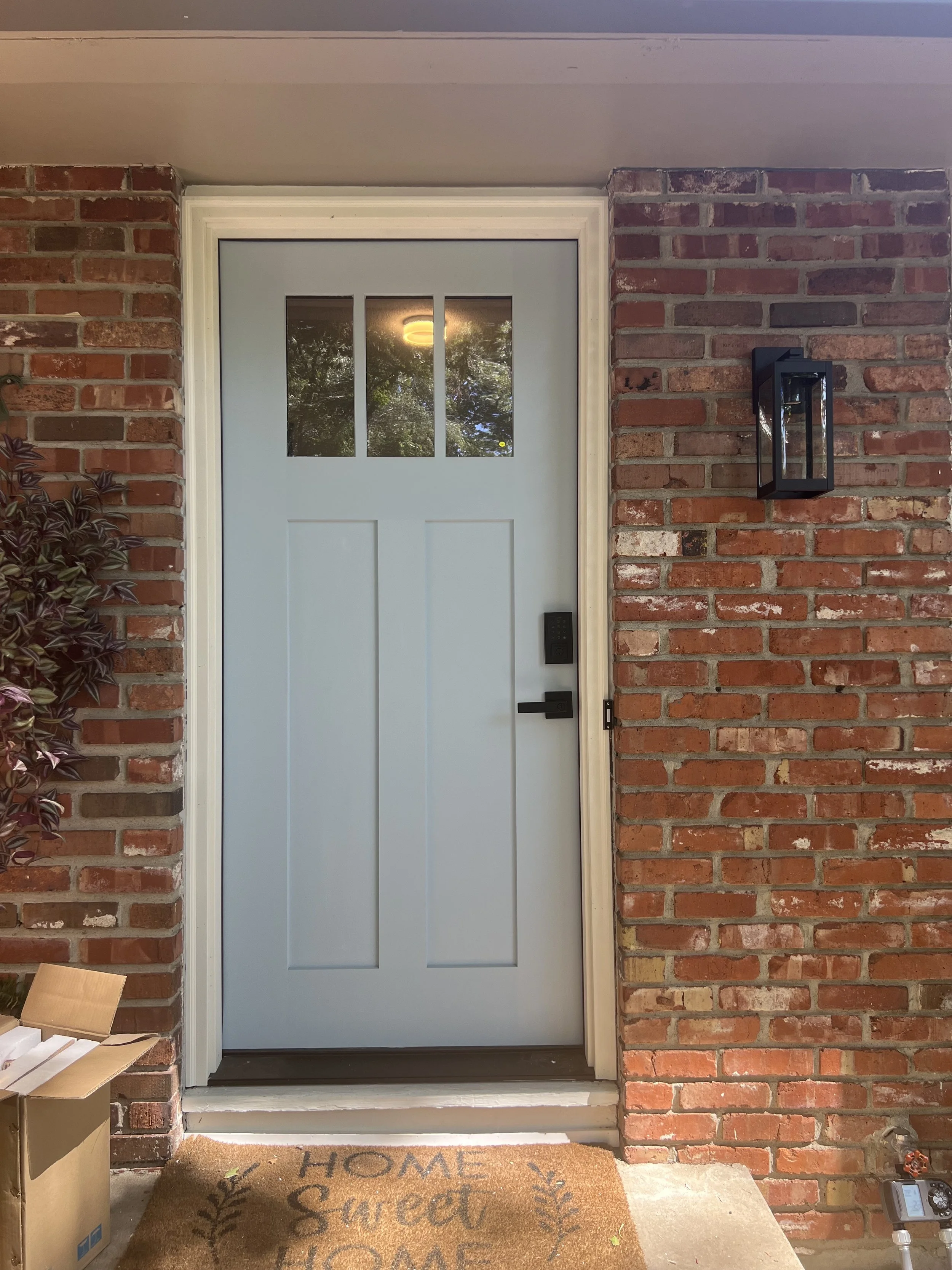 Light blue front door with three small windows at the top, set in a brick wall. A black outdoor lantern-style light fixture is mounted to the right of the door. A welcome mat with the words "Home Sweet Home" is on the ground in front of the door, with a cardboard box on the left and a small electronic device on the right.