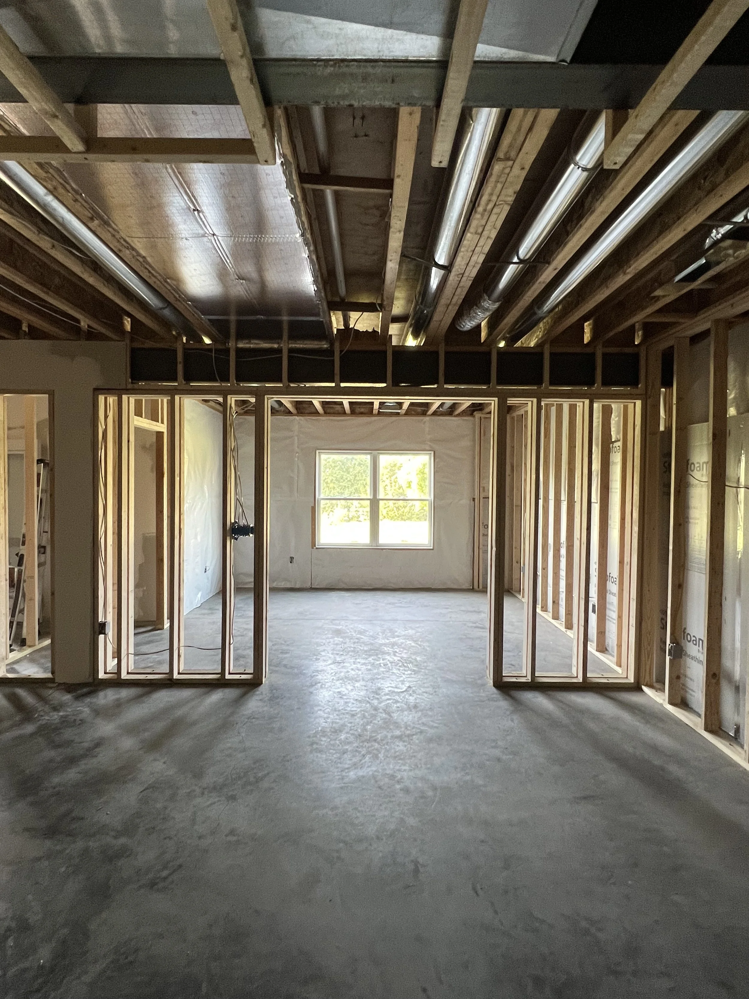 Interior of a house under construction, showing framed walls and ceiling with exposed wooden beams, pipes, and insulation, and a window at the far end letting in natural light.