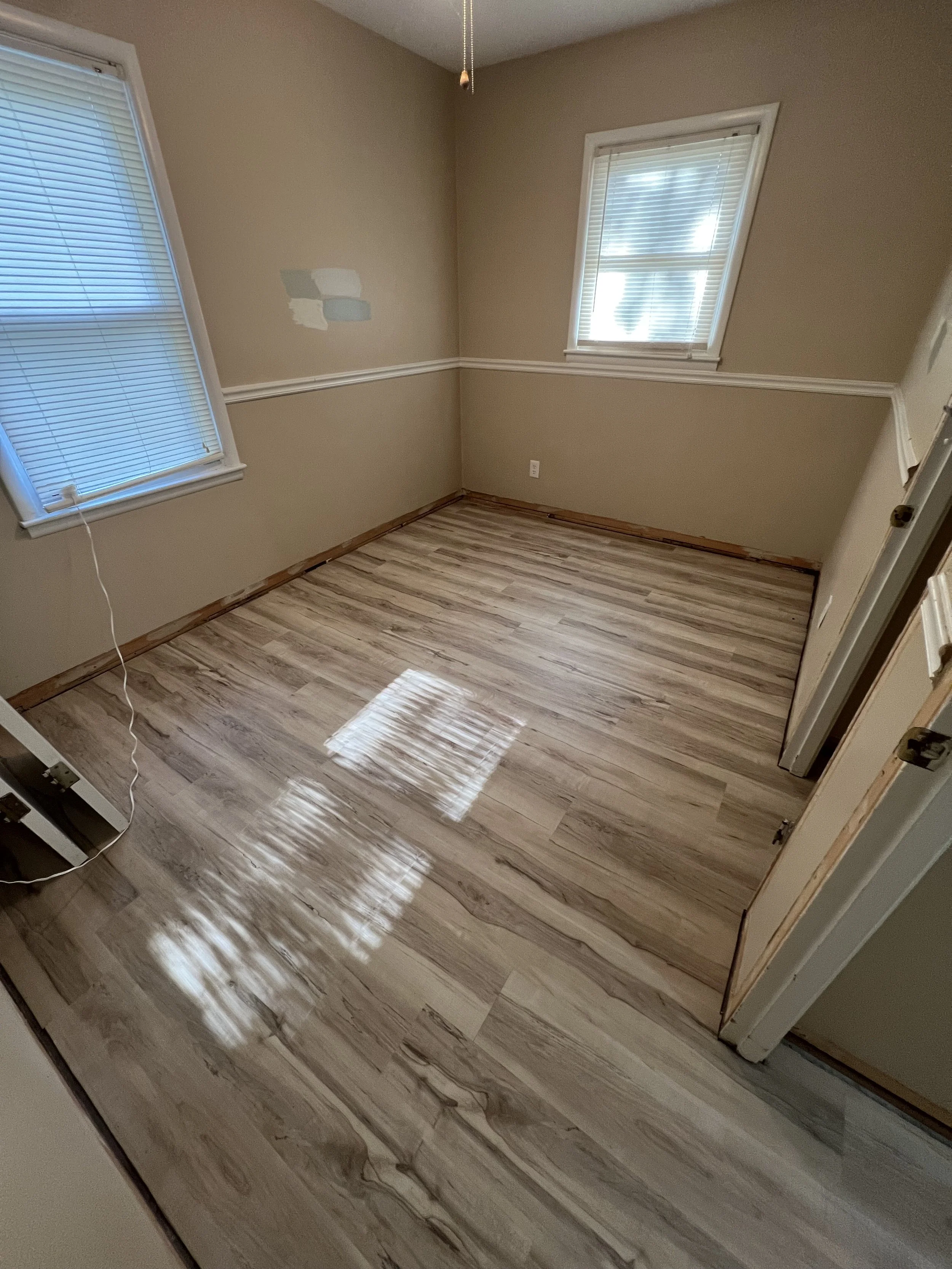 Empty room with newly installed wood laminate flooring, beige walls, two windows with blinds, and a ceiling fan pull chain.