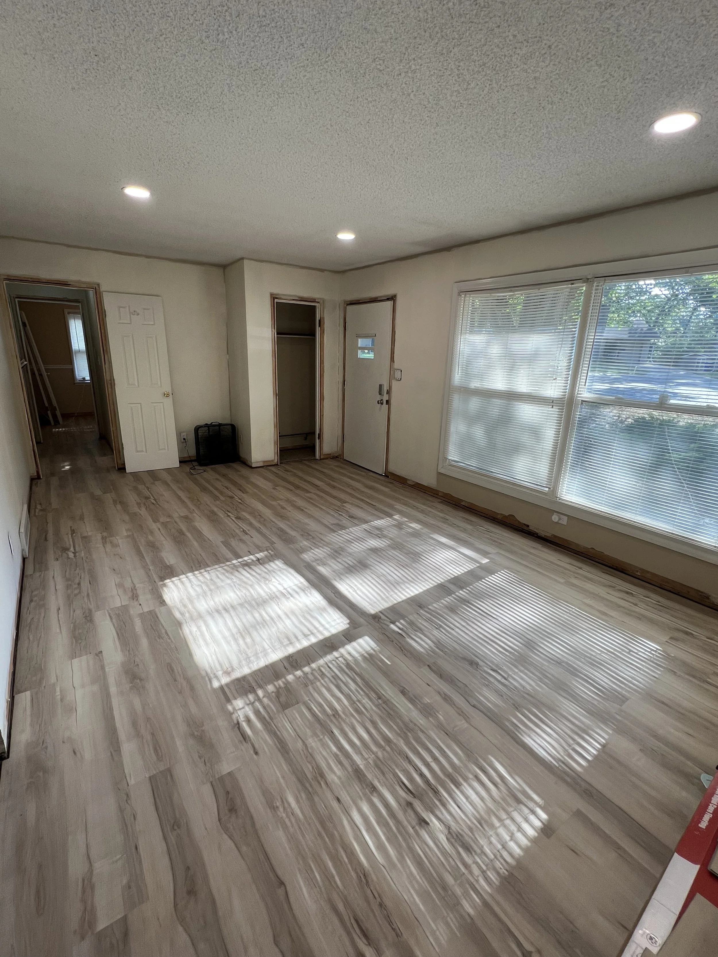 Empty living room with light wood flooring, large window with blinds, white walls, and ceiling lights.
