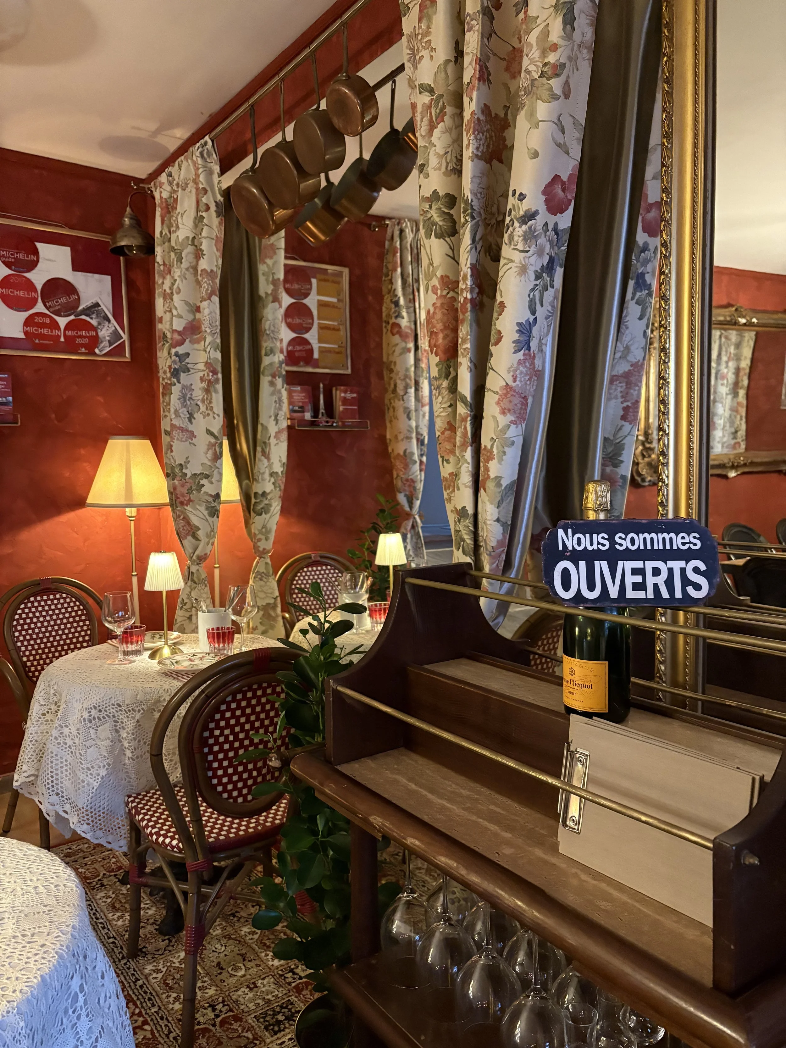 Interior of a cozy restaurant with a table set with glassware and a small lamp, surrounded by floral curtains, wooden chairs with red and white patterned seats, and a mirror reflecting part of the room. A sign on a bar reads "Nous sommes OUVERTS" ind
