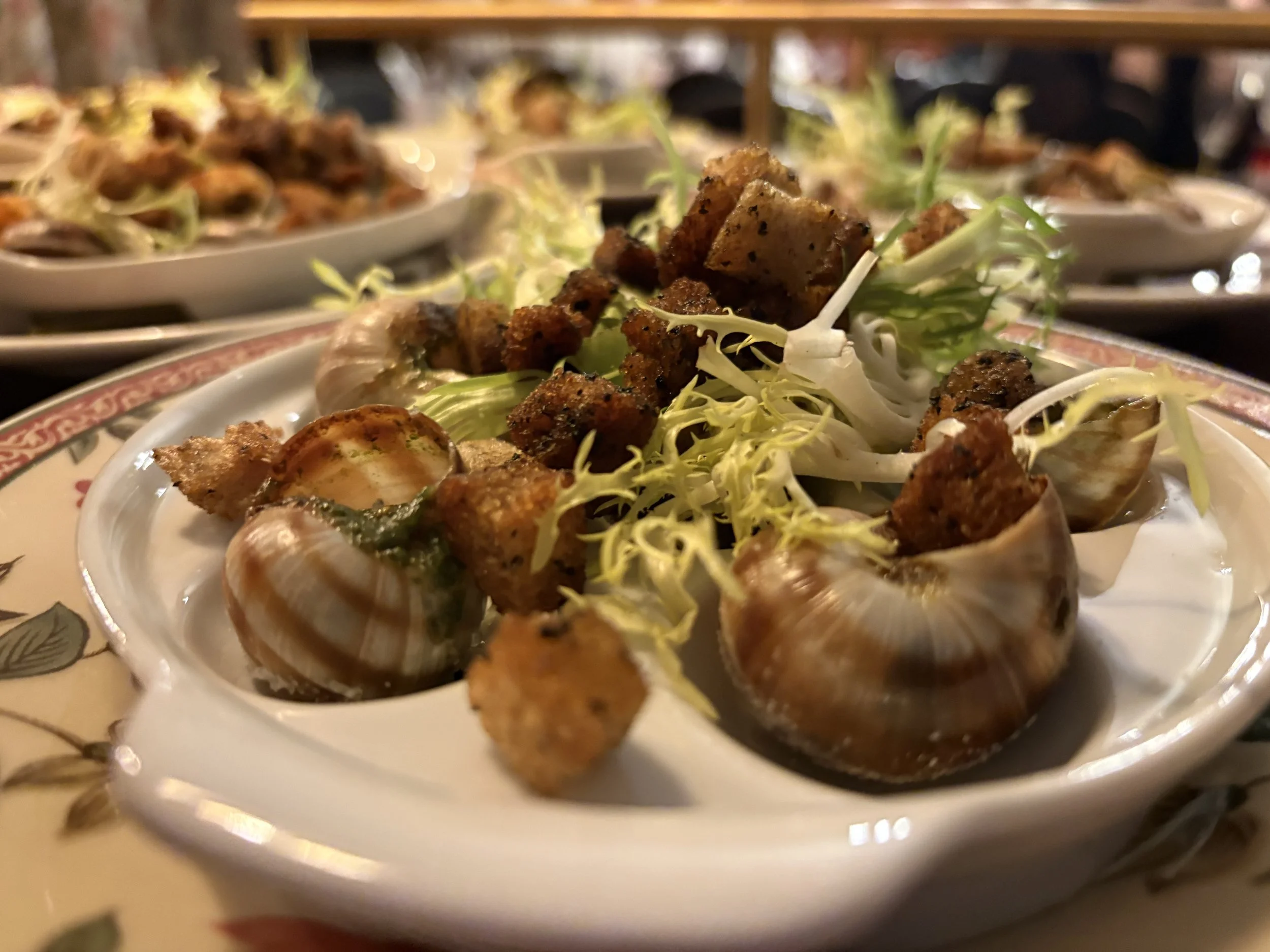 Close-up of a seafood dish with clams, fried breaded meat or fish cubes, and shredded lettuce on a decorative plate, with more plates of similar food in the background.