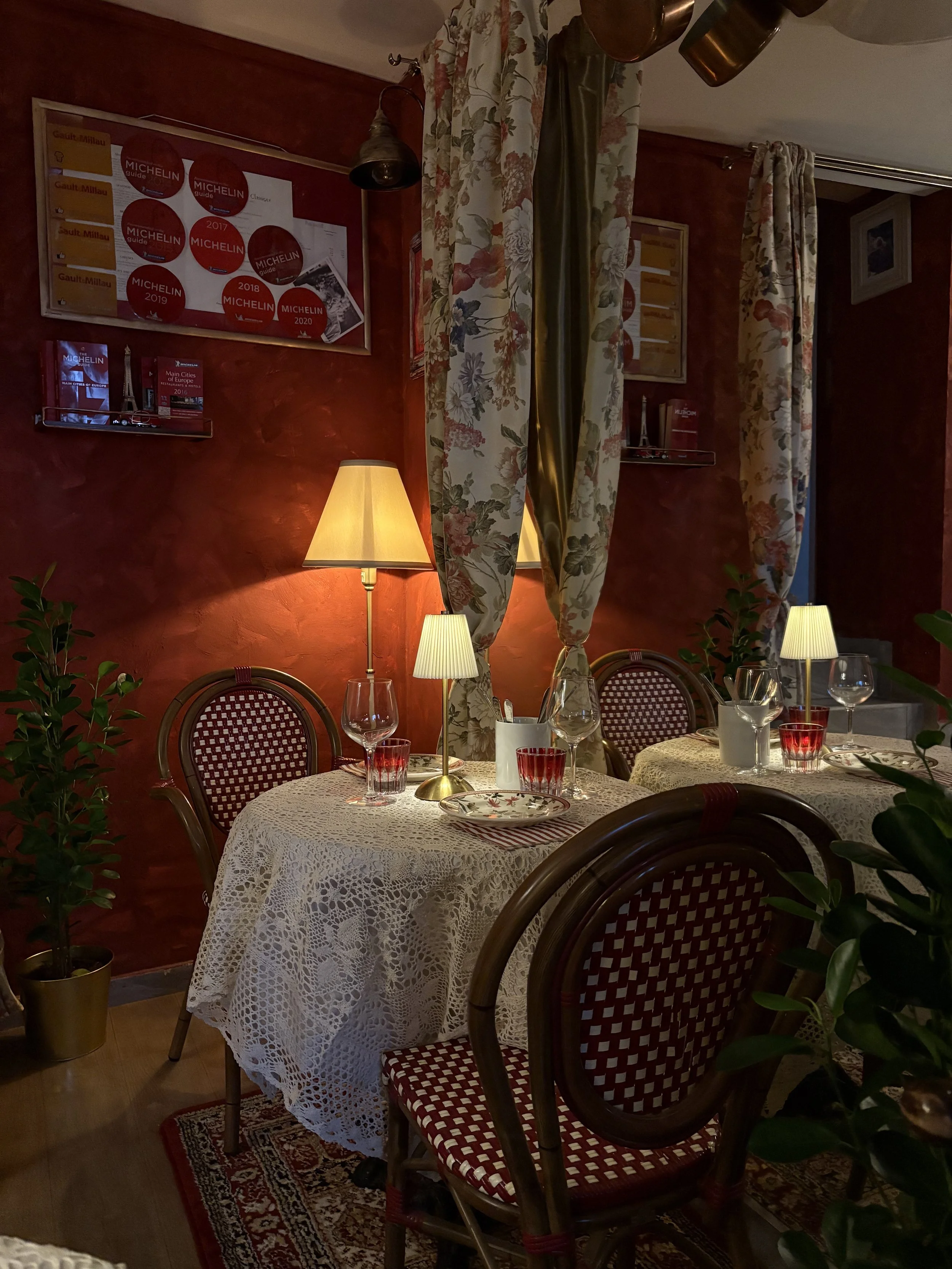 A cozy restaurant setting with round tables covered in lace tablecloths, surrounded by wooden chairs with woven red and white seats. There are potted plants, small table lamps, wine glasses, and dinnerware on the tables. The warm-colored walls are de