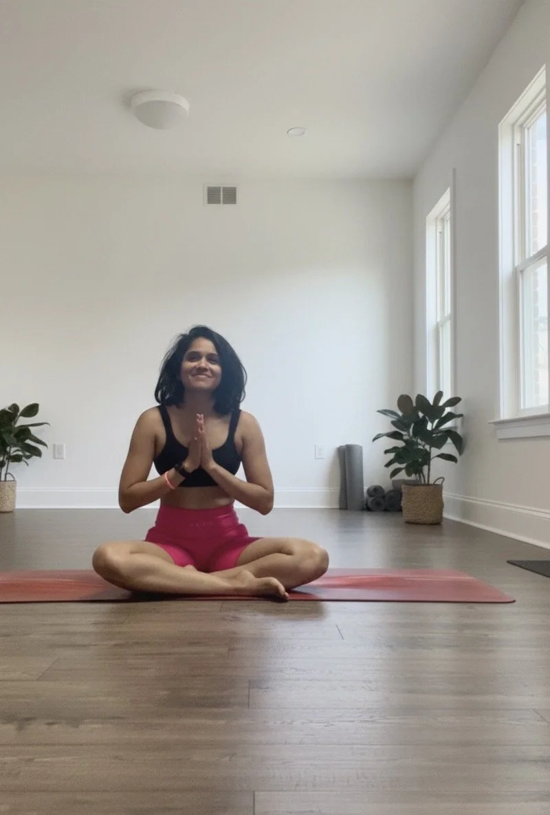 A woman sitting cross-legged on a yoga mat in a bright, minimalistic room, practicing yoga with her hands in a prayer position.
