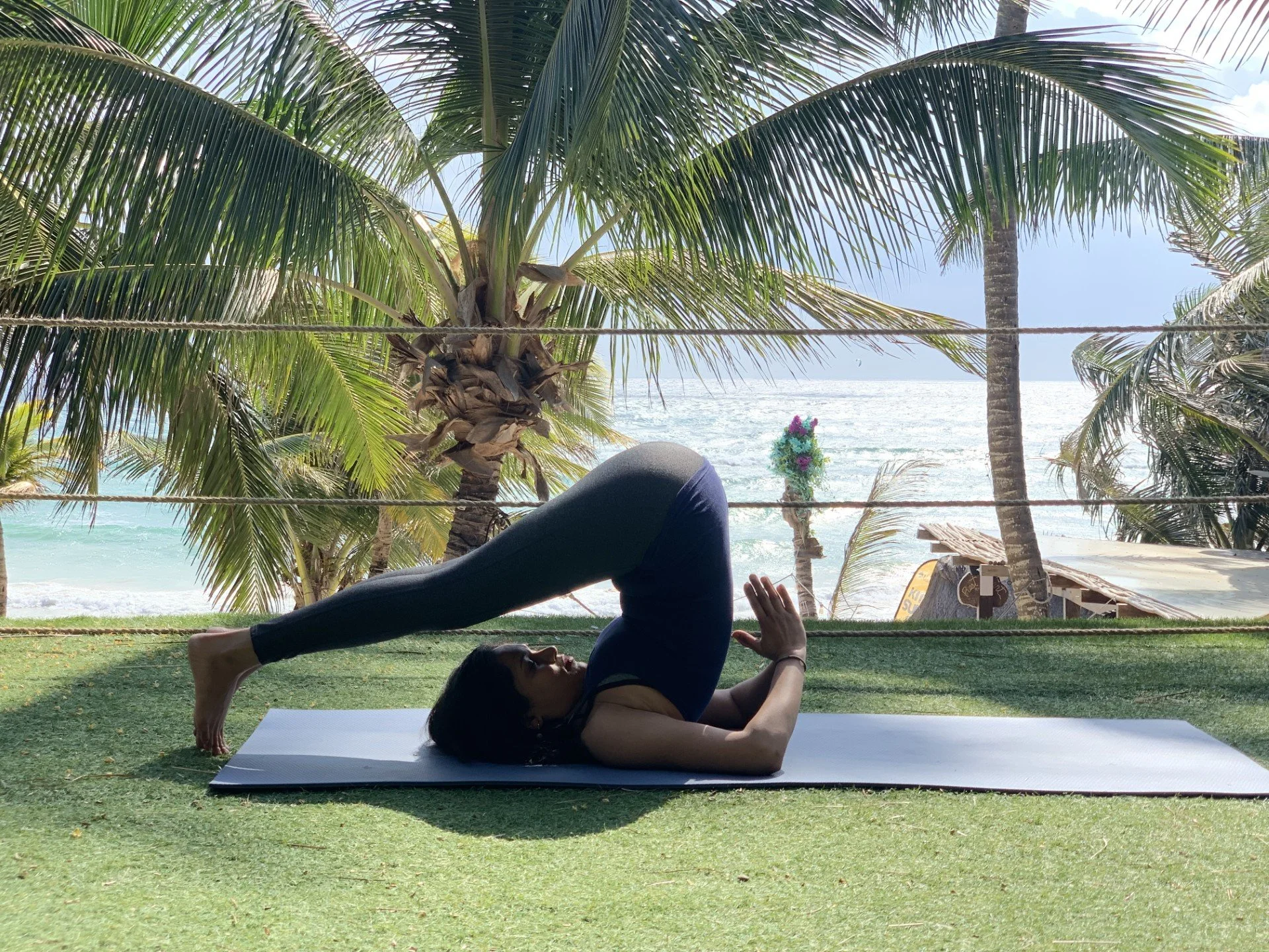 A woman practicing yoga on a mat outdoors near the beach, with palm trees and ocean in the background.