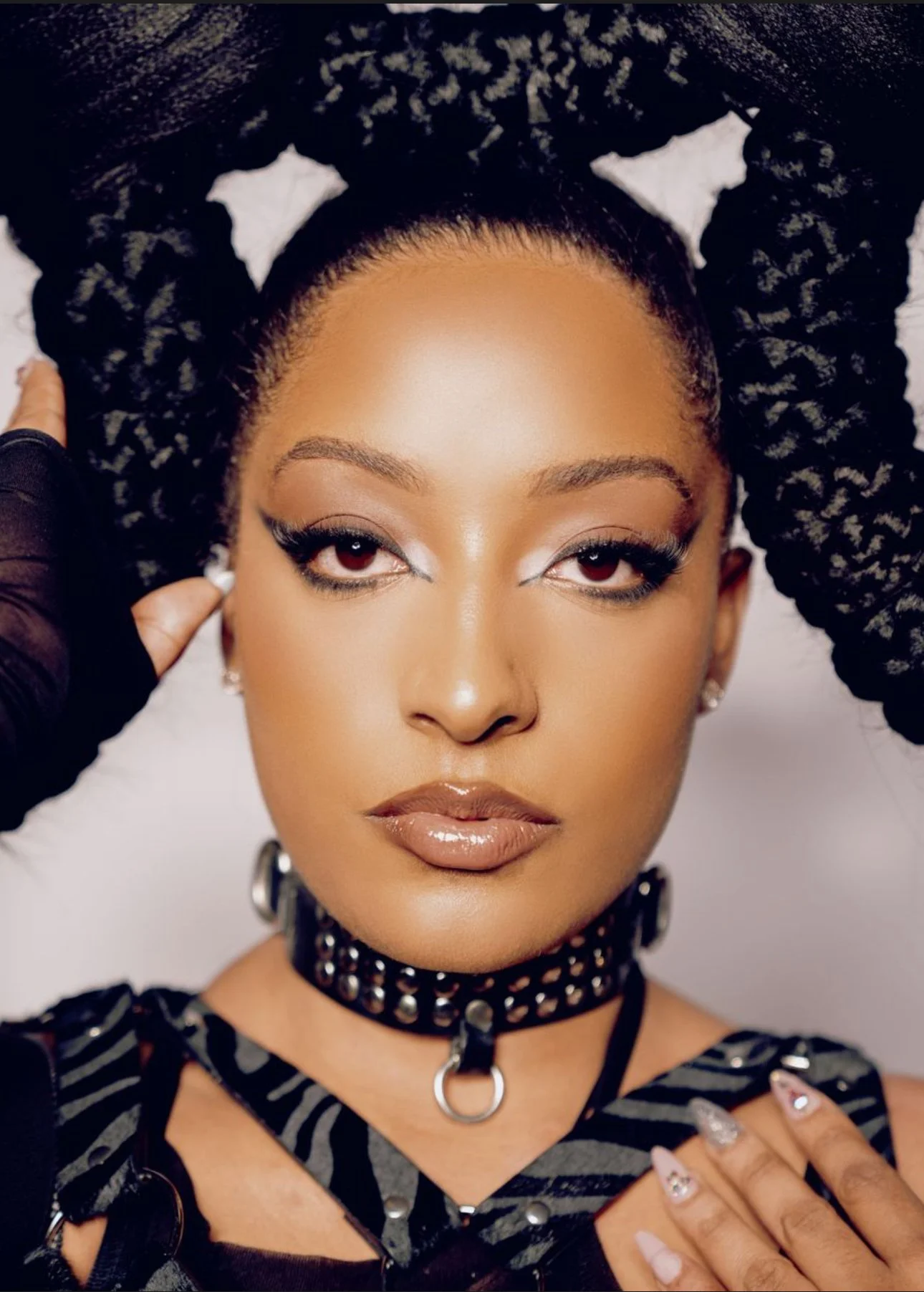 Portrait of a woman with styled hair, makeup, and accessories, including a choker necklace and earrings, against a light background.