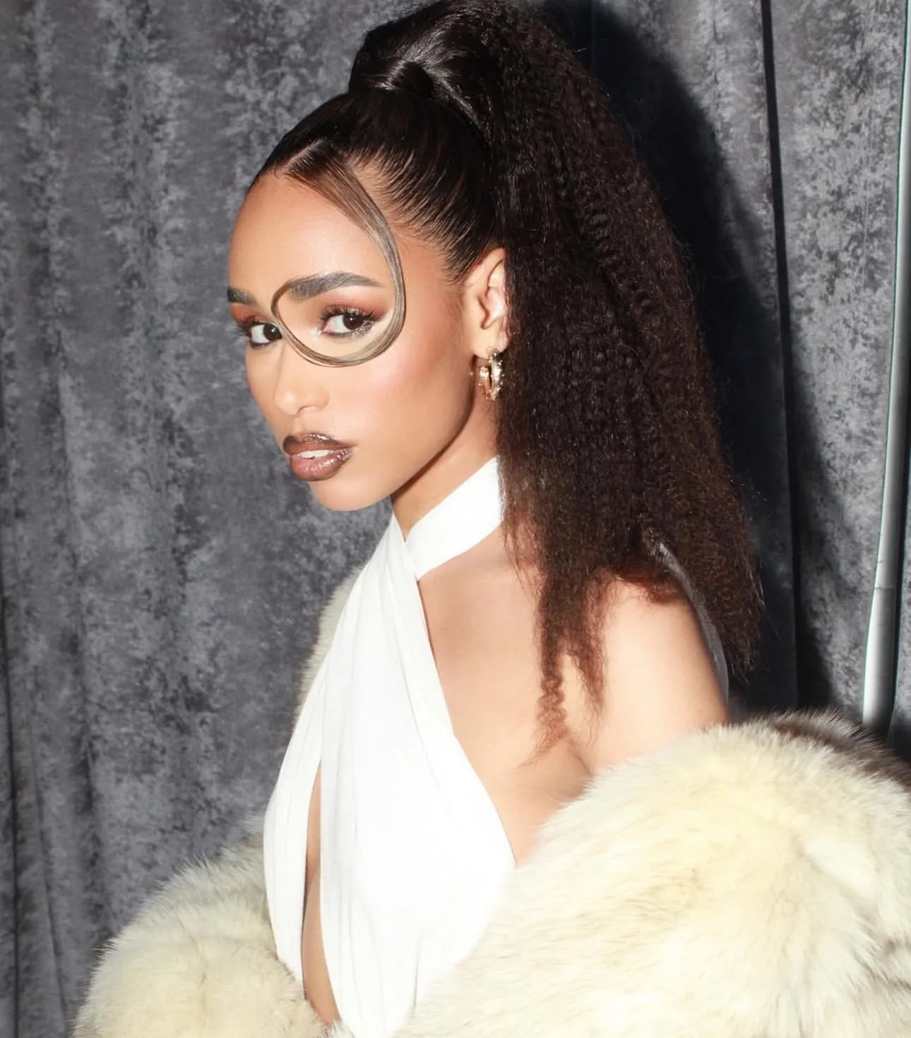 A woman with a distinctive hairstyle, wearing a white dress and a fur stole, posing against a textured background.
