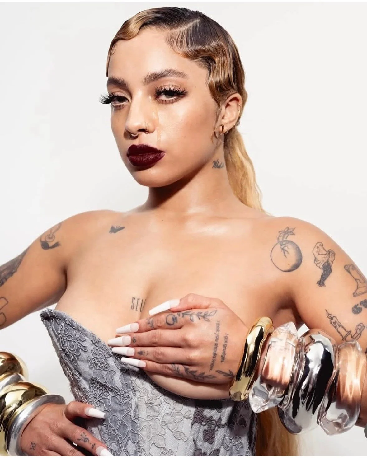 A woman with multiple tattoos and large bracelets poses with her hand over her chest, wearing dark lipstick and eye makeup, against a plain white background.