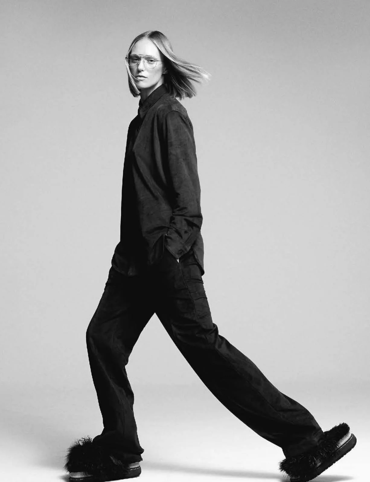 Black and white photo of a woman with short hair and glasses walking with her hands in her pockets, wearing a black shirt, black pants, and fluffy black shoes, against a plain background.