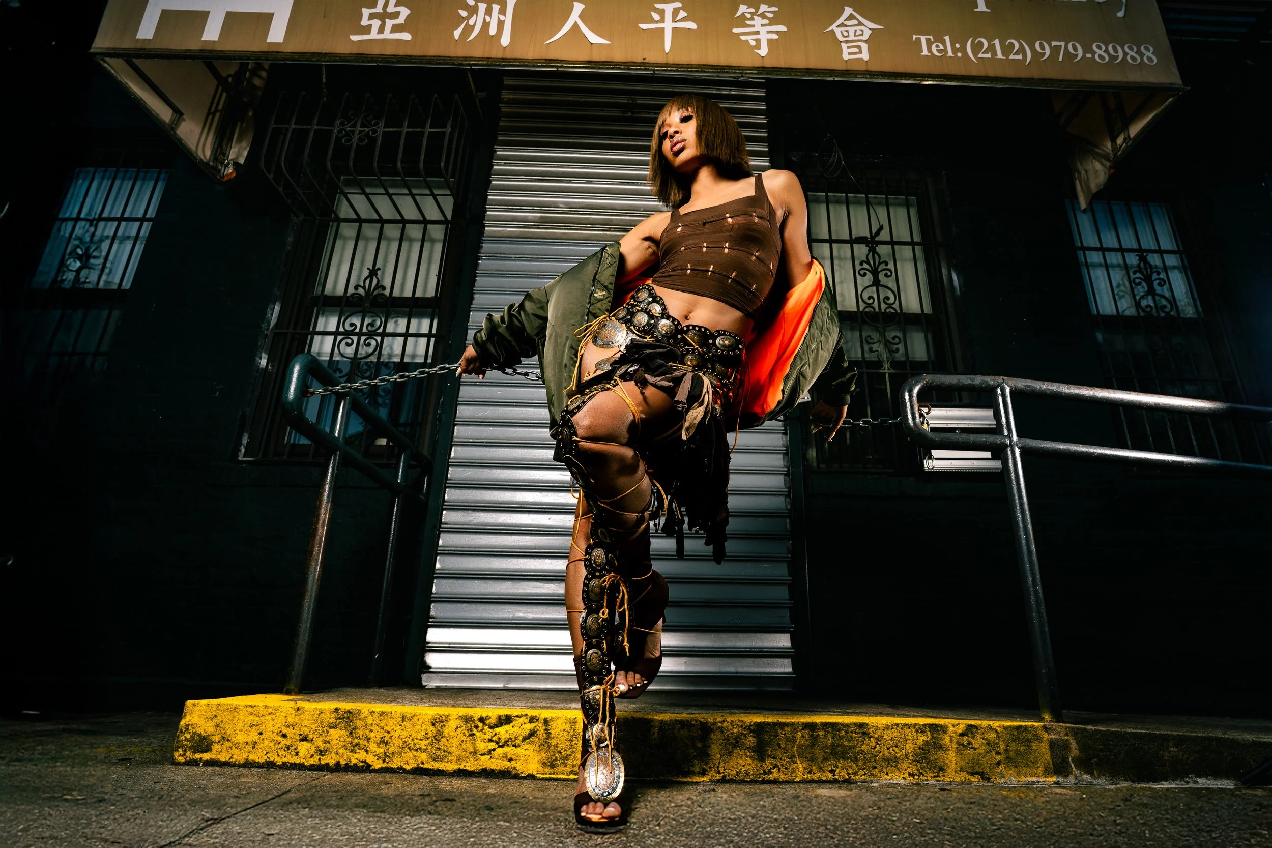 Stylish woman in edgy outfit standing in front of a closed shop with Chinese characters, metal shutter, and barred windows, posing with one leg raised and wearing high-heeled sandals with decorative embellishments.