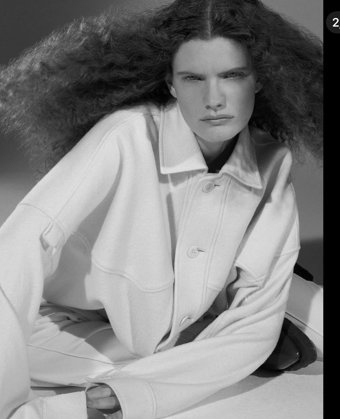 Black-and-white portrait of a woman with voluminous, curly hair, wearing a light-colored jacket, looking directly at the camera.