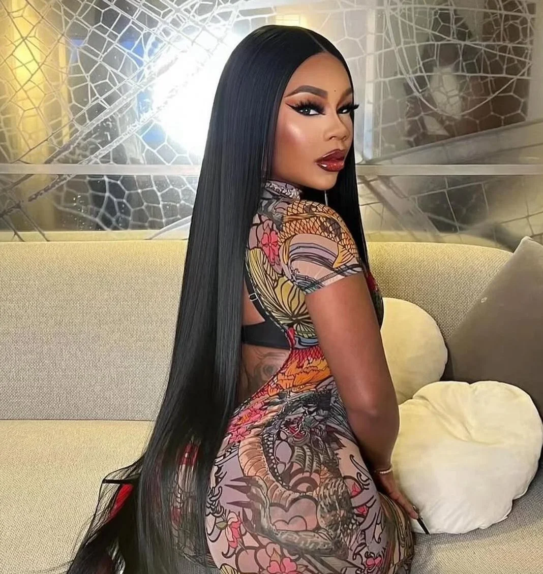 A woman with long straight black hair, wearing a colorful tattoo-style dress, sitting on a beige couch with patterned backdrop.