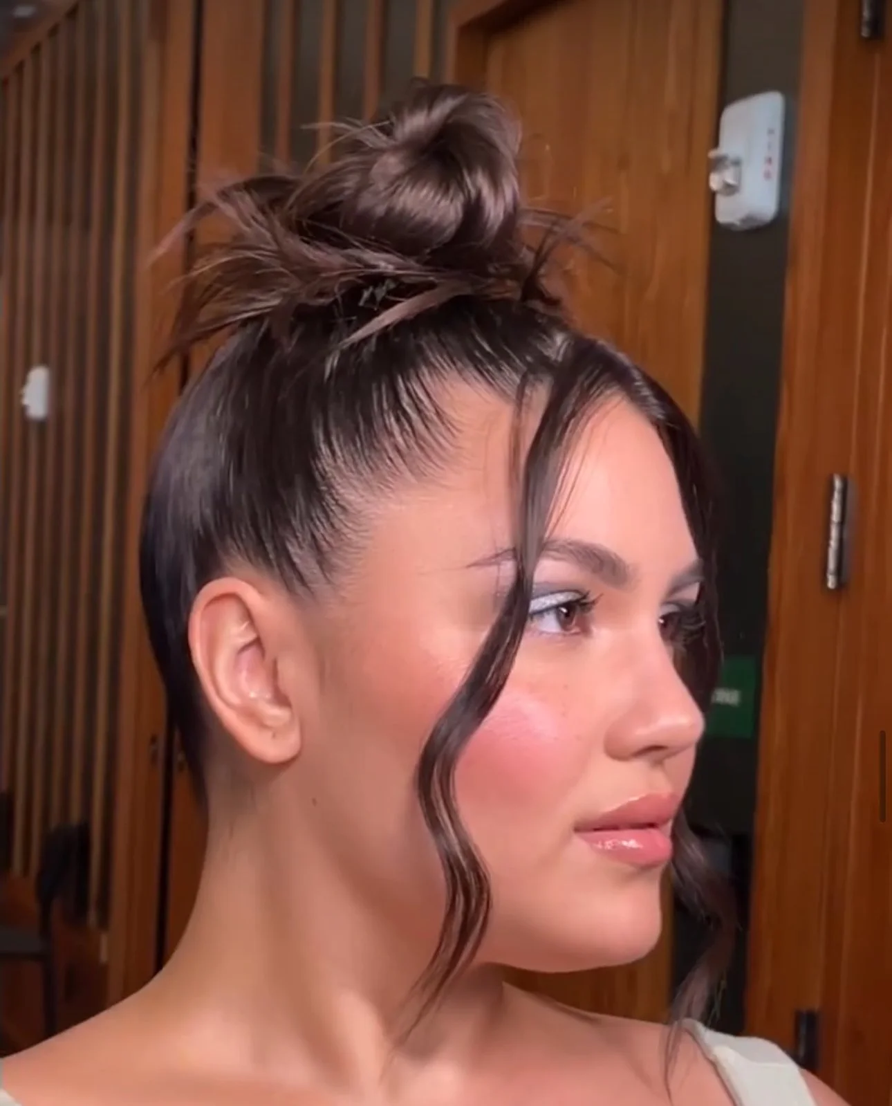 A woman with styled dark brown hair in a high bun with loose curls and face-framing strands, posing indoors.