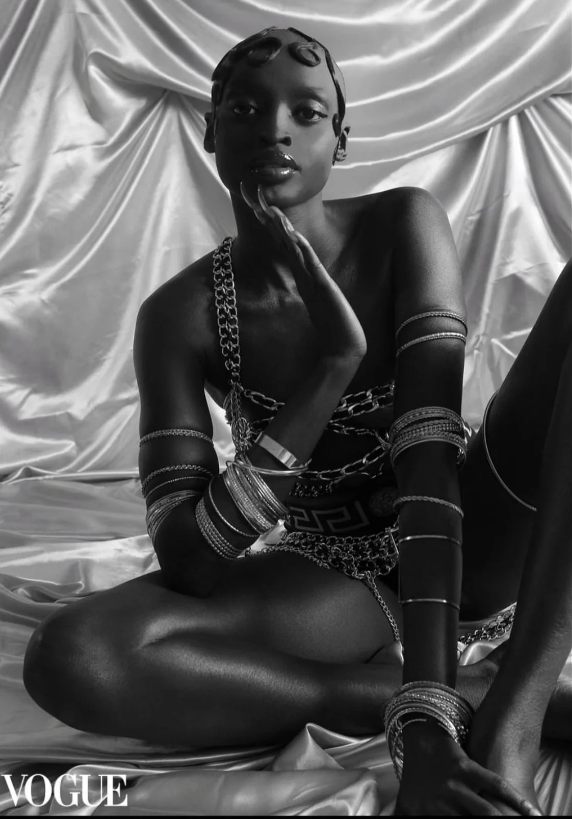 Black and white photo of a woman sitting on the floor surrounded by silky fabric, wearing multiple jewelry pieces including bracelets, armlets, and a chain, with a confident expression and stylish pose.